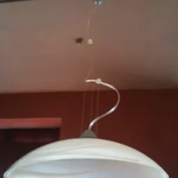 Biała lampa wisząca z kloszem w kształcie talerza, zawieszona na przezroczystym kablu, widoczna na tle białego sufitu i bordowej ściany.