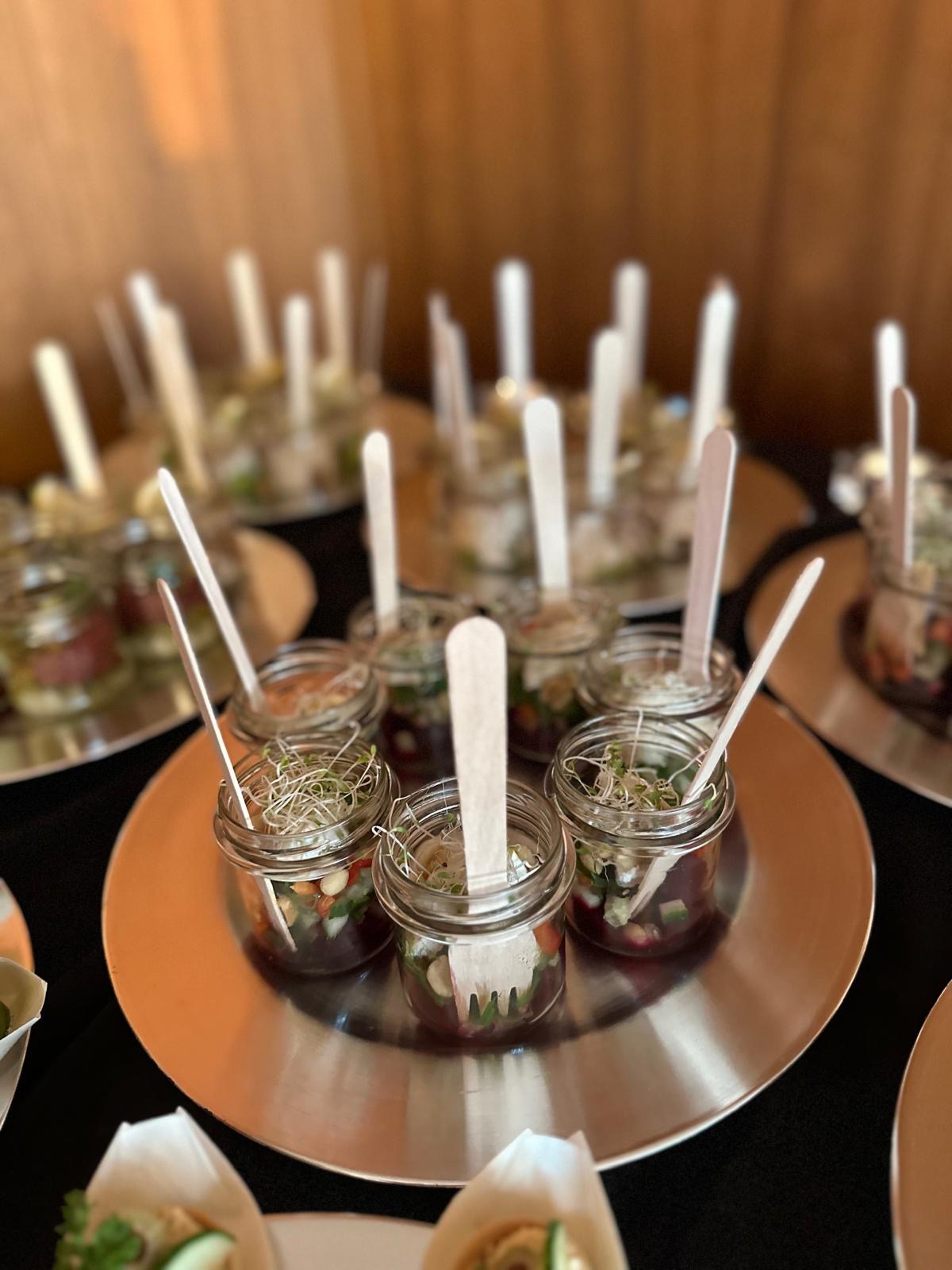 Szklane słoiczki z sałatką i drewnianymi widelczykami na srebrnej tacy, część cateringu na ciemnym stole, w tle kolejne porcje.