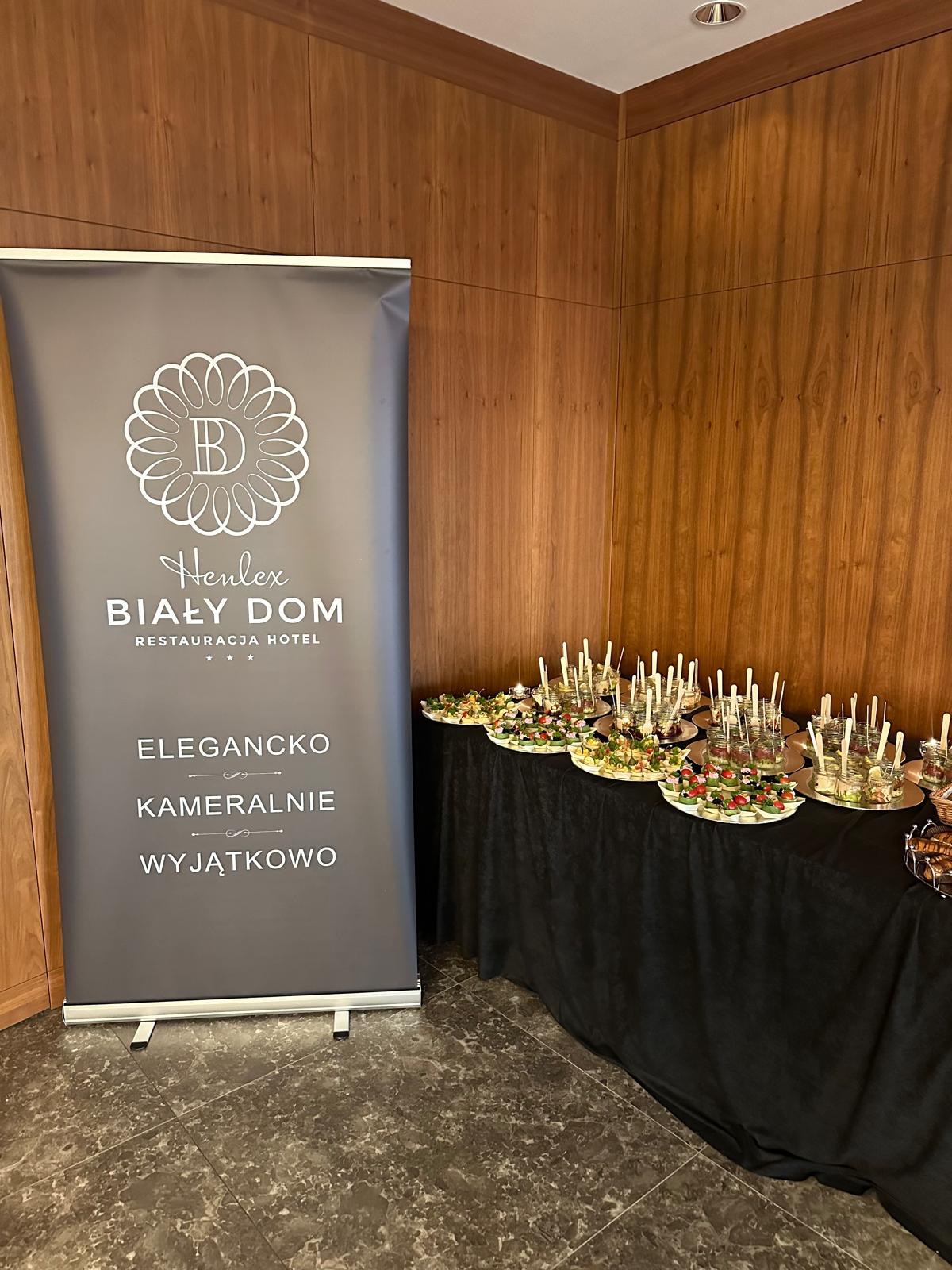 Stół cateringowy z przekąskami obok roll-upu reklamującego restaurację Henlex Biały Dom, elegancko, kameralnie, wyjątkowo. Drewniane panele na ścianie.