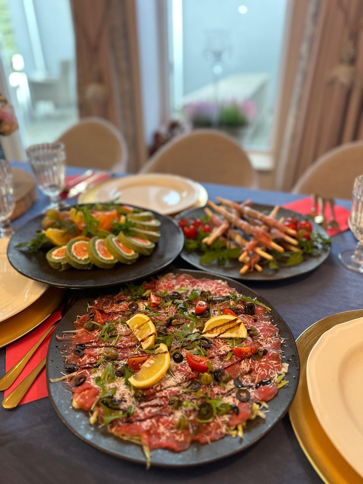 Elegancko zastawiony stół z trzema talerzami wykwintnych przekąsek: carpaccio, roladki z łososiem i szaszłyki z szynki parmeńskiej, gotowy na przyjęcie.