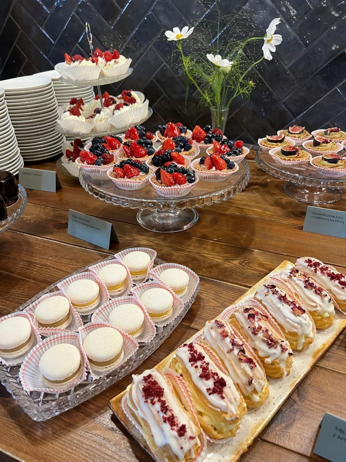 Elegancki stół z deserami: tartaletki z owocami, makaroniki i eklerki z kremem, idealne na przyjęcie. W tle ciemna ściana i białe kwiaty w wazonie.