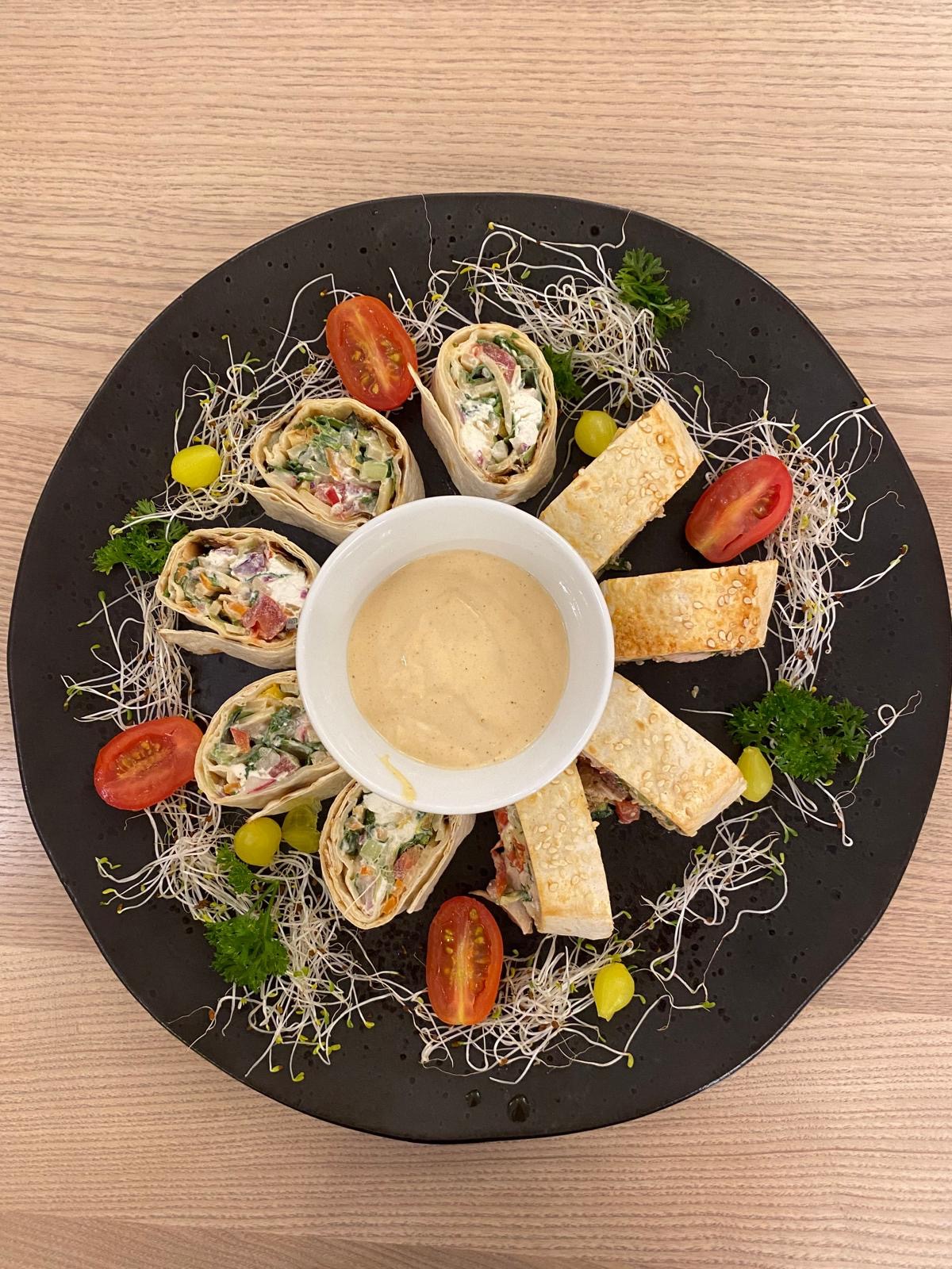 Czarny talerz z roladkami tortilli z warzywami i sosem, udekorowany kiełkami, pomidorkami cherry i zielonymi winogronami. Elegancka kompozycja cateringowa.