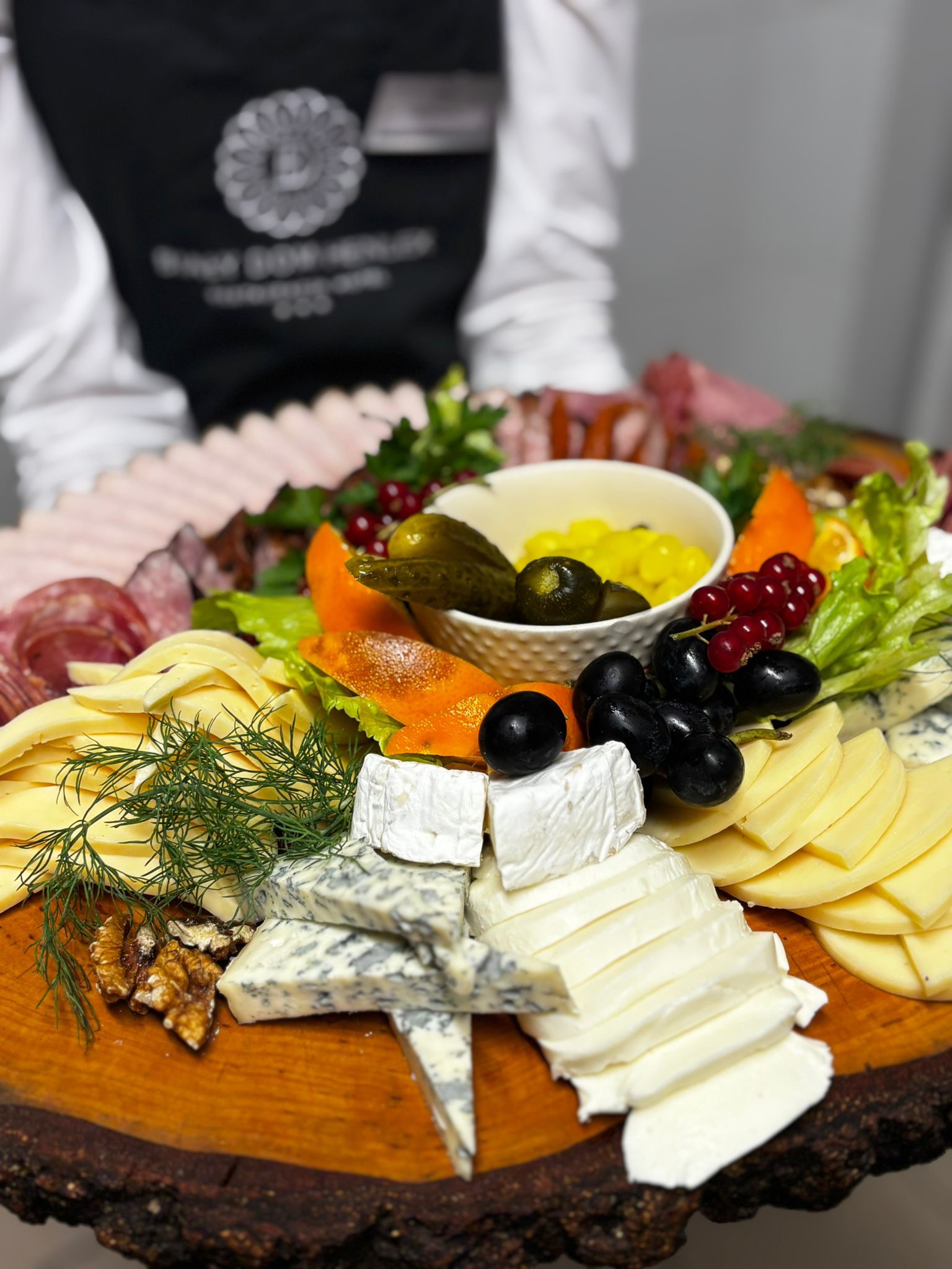 Deska serów i wędlin na drewnianym plastrze, z dodatkami owoców, warzyw i marynat, prezentowana przez osobę w fartuchu. Elegancki catering.