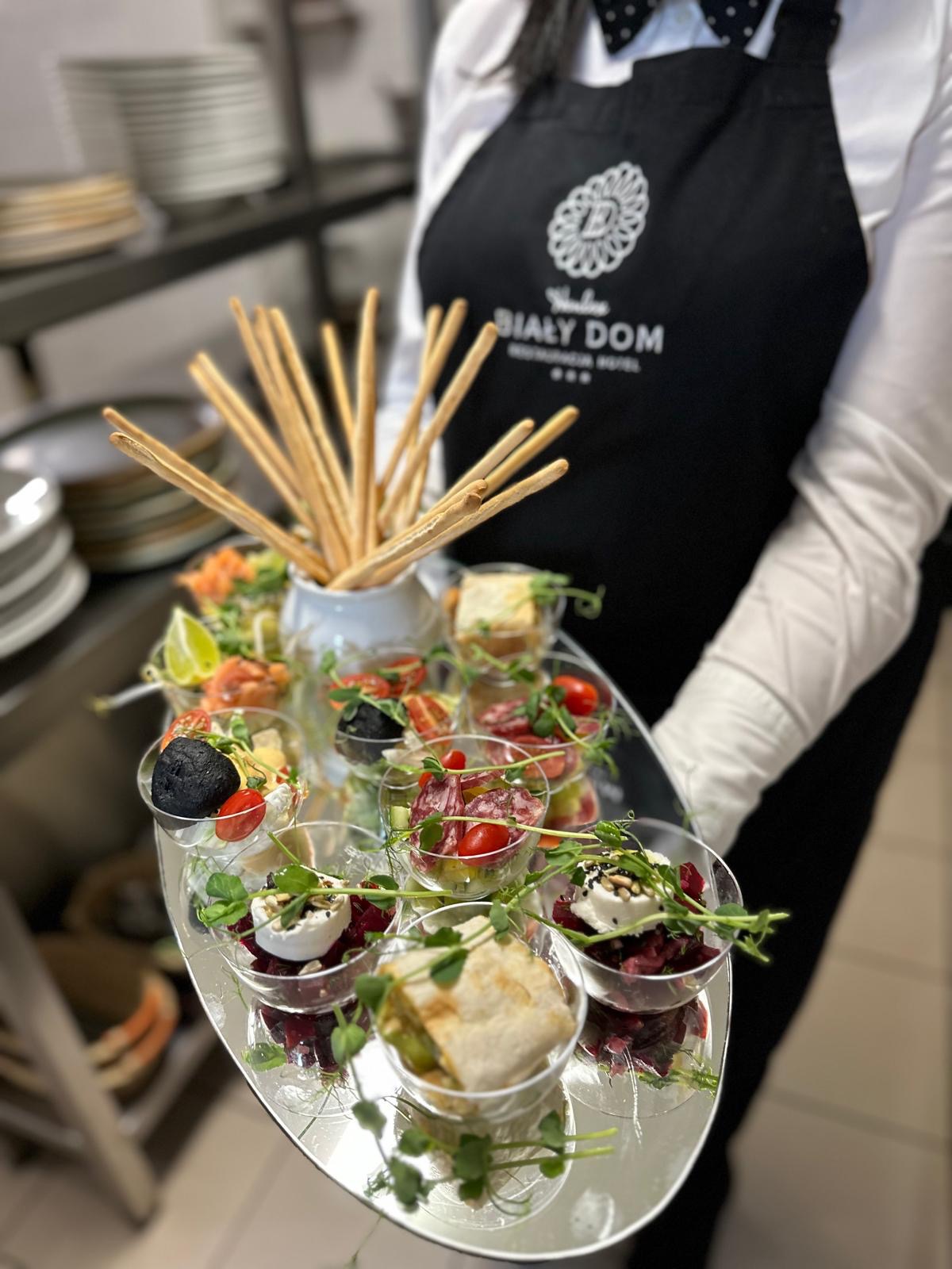 Kelner w firmowym fartuchu prezentuje tacę z wykwintnymi przekąskami w szklanych naczyniach, w tle zastawa stołowa i elementy wyposażenia kuchni.