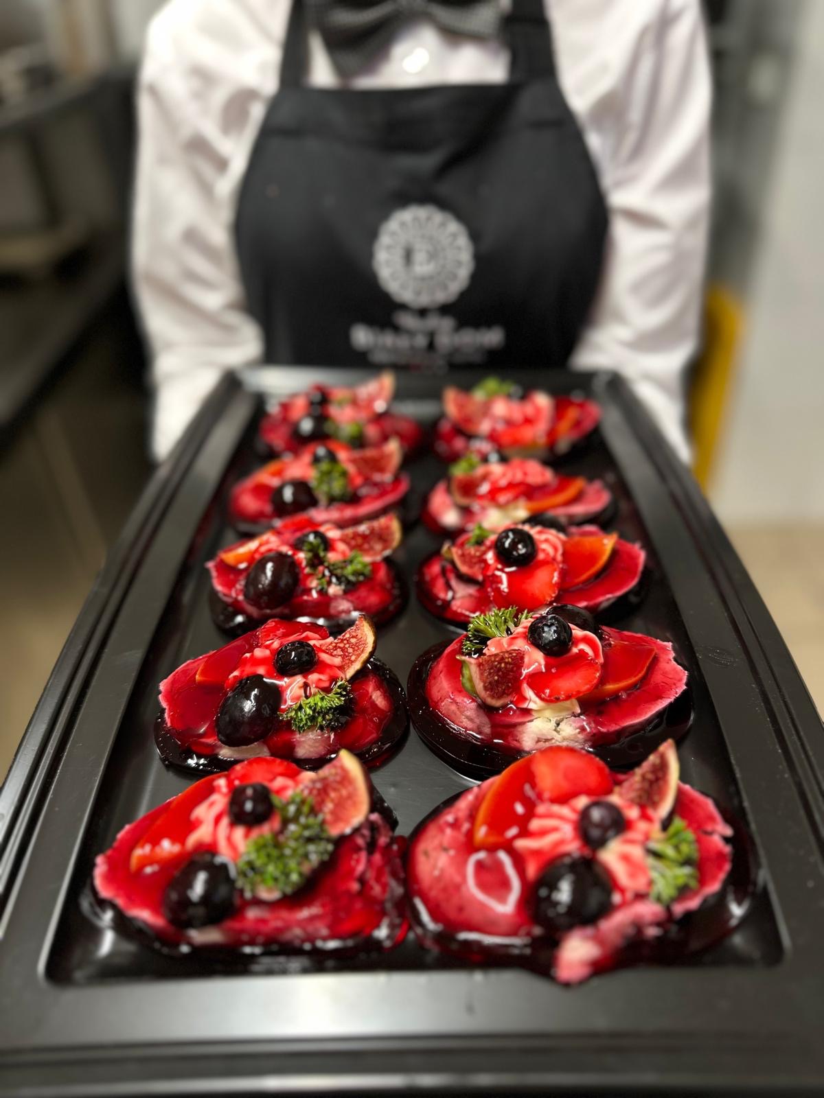 Kelner w uniformie prezentuje tacę z wykwintnymi deserami: galaretki z figami, winogronami i ziołami, idealne na elegancki catering.