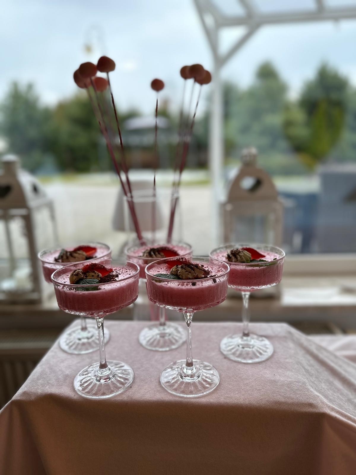 Elegancko podane desery w szklanych pucharkach na tle okna z widokiem na zieleń. Dekoracja stołu z kwiatami i lampionami. Słodki poczęstunek na przyjęciu.