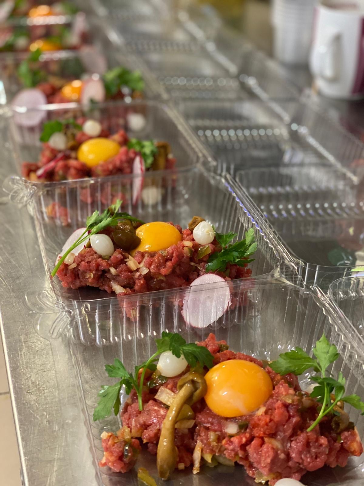 Tatar wołowy z żółtkiem, rzodkiewką i marynatami w przezroczystych pojemnikach, gotowy do transportu. Catering na metalowym blacie.