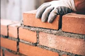 Mur z czerwonej cegły w trakcie wznoszenia, dłoń w rękawicy murarskiej układa kolejną cegłę. Widoczna zaprawa i fragment muru. Ujęcie z bliska.