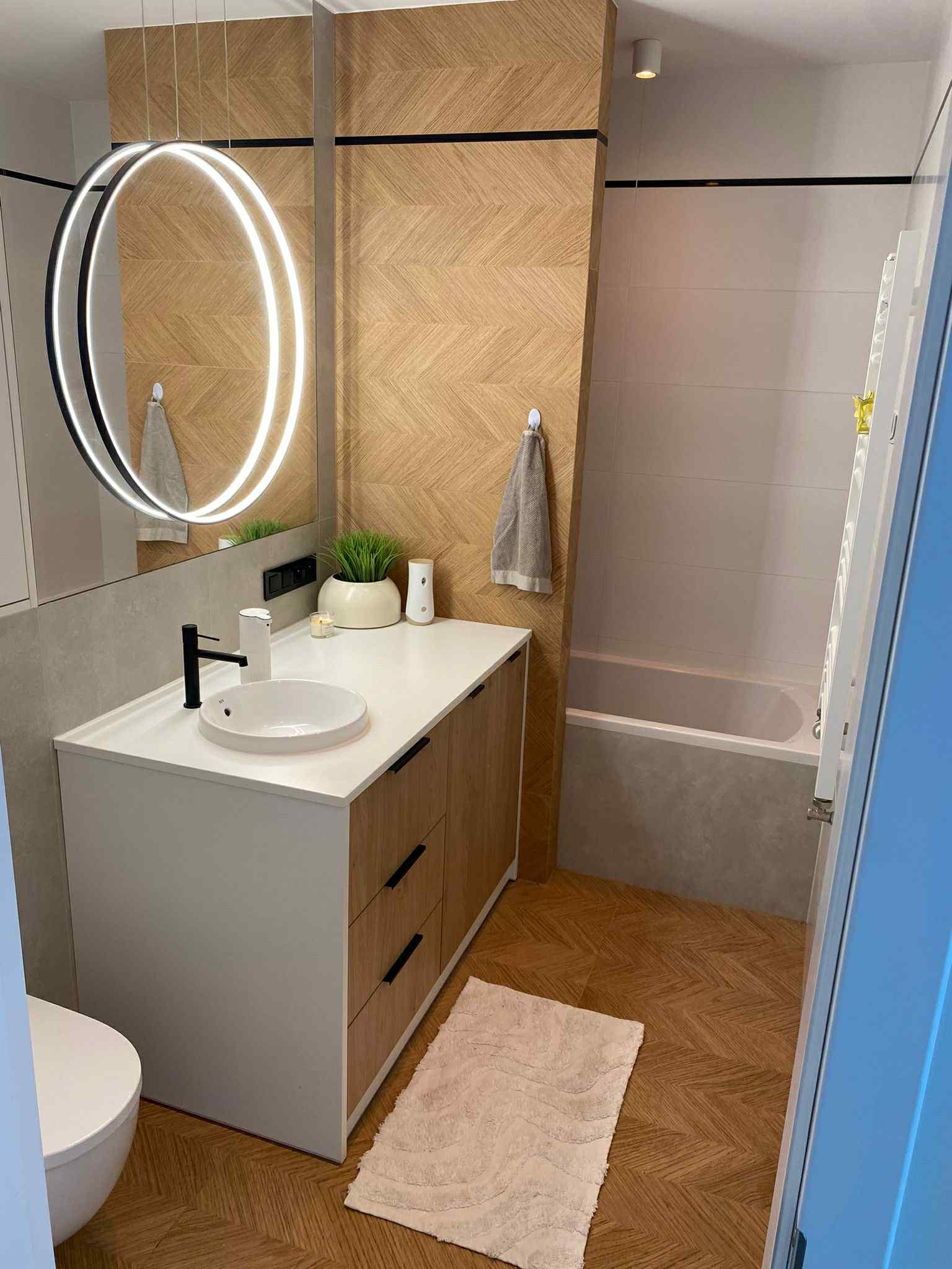 Nowoczesna łazienka z drewnianą mozaiką na ścianie, okrągłym lustrem z podświetleniem LED i białymi meblami łazienkowymi z drewnianymi akcentami.