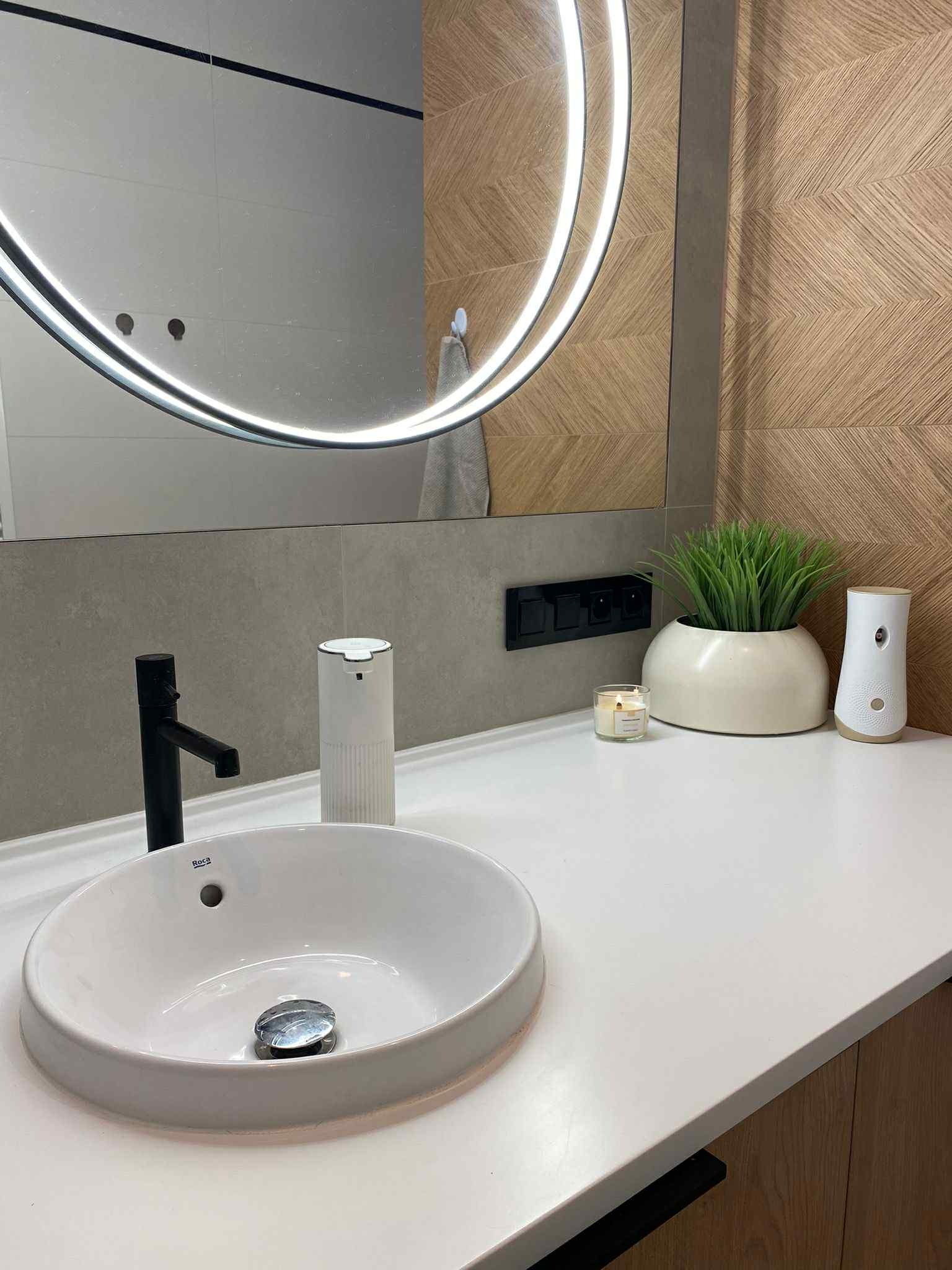 Nowoczesna łazienka: owalne lustro z podświetleniem LED, biała umywalka Roca z czarną armaturą, drewniana mozaika na ścianie i dekoracje.