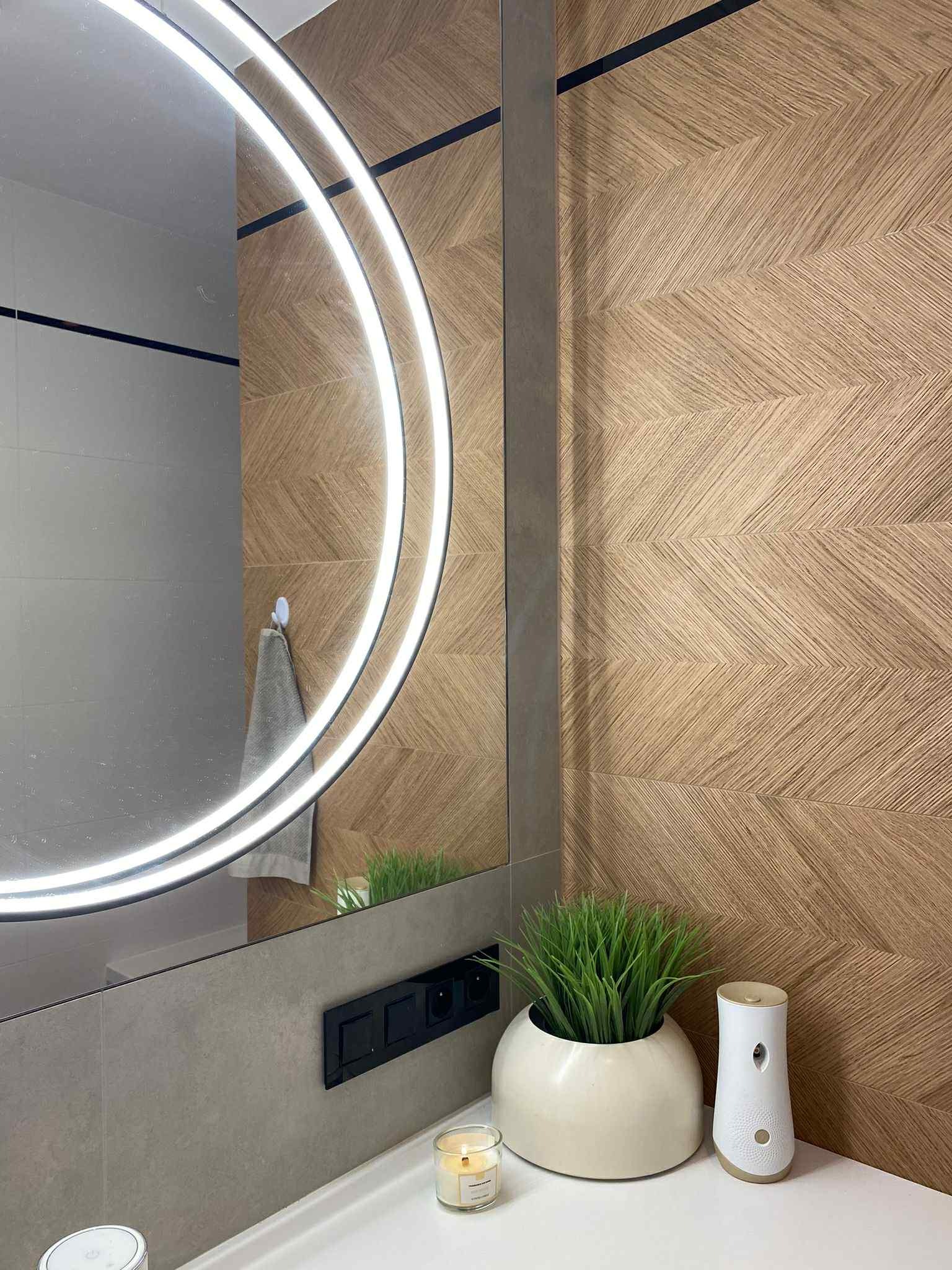 Nowoczesna łazienka z okrągłym lustrem LED, drewnopodobnymi płytkami ułożonymi w jodełkę i minimalistycznymi dodatkami na blacie.