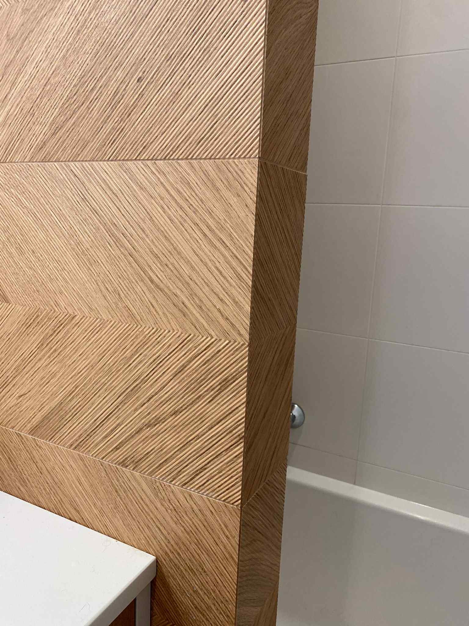 Drewnopodobne płytki ułożone w jodełkę na ścianie w łazience, obok białe płytki i fragment wanny. Dekoracyjne wykończenie z bliska.