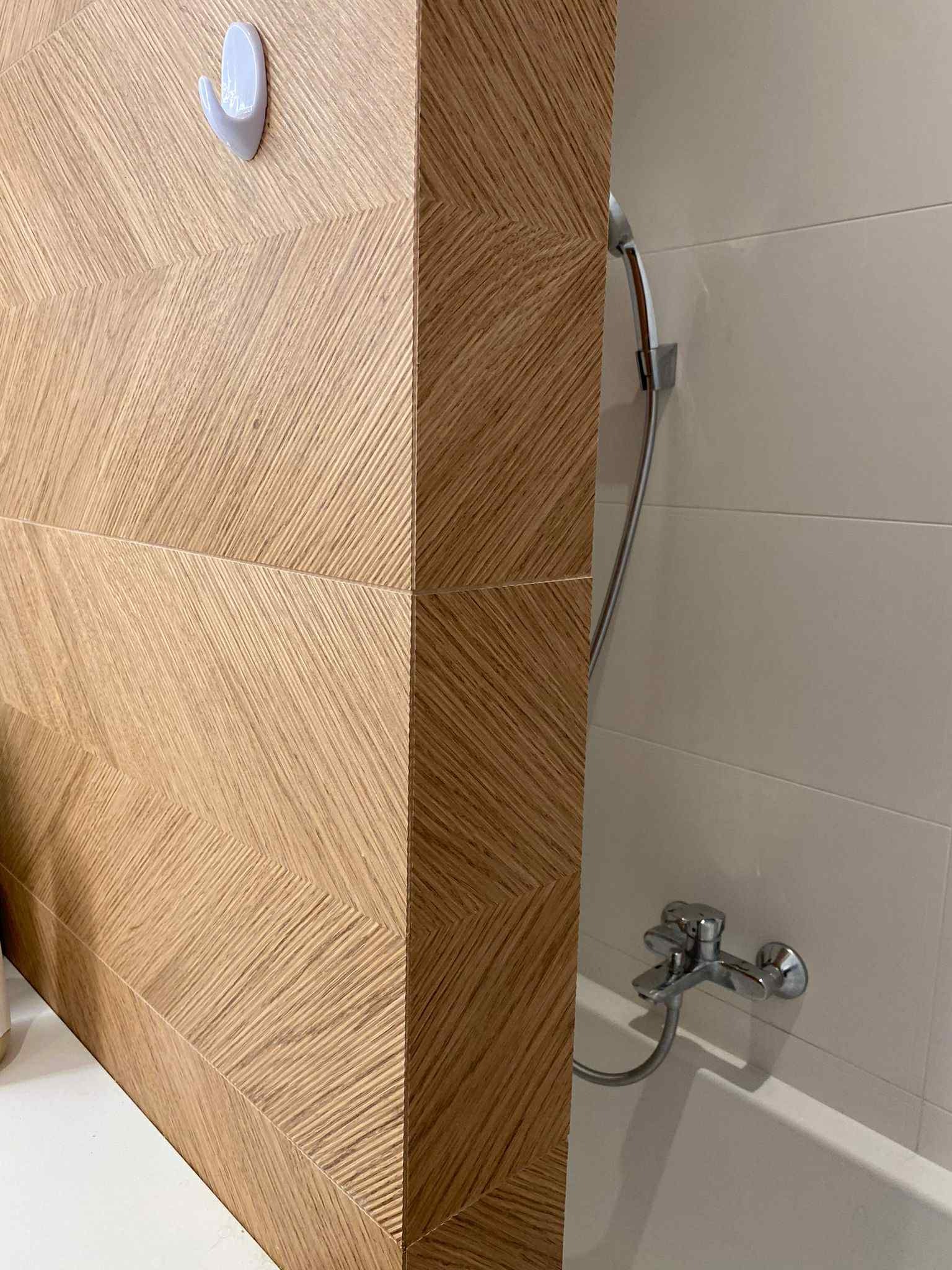 Wykończona łazienka: drewnopodobne płytki ułożone w jodełkę na ścianie, chromowana bateria wannowa i biały haczyk na ręcznik. Nowoczesny design.