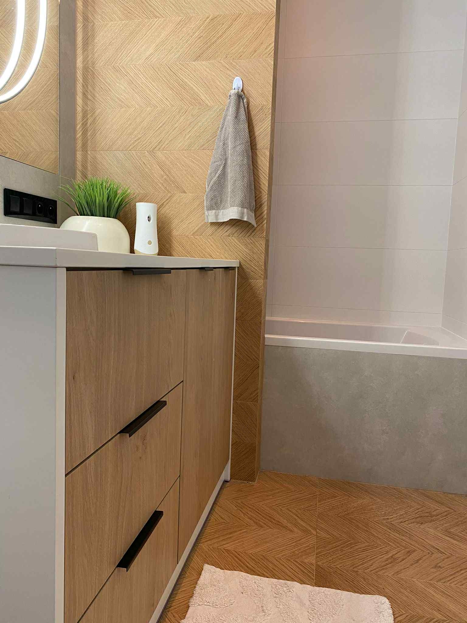 Nowoczesna łazienka z drewnopodobnymi płytkami ułożonymi w jodełkę na ścianie i podłodze. Szafka pod umywalką z drewnianymi frontami i czarnymi uchwytami.