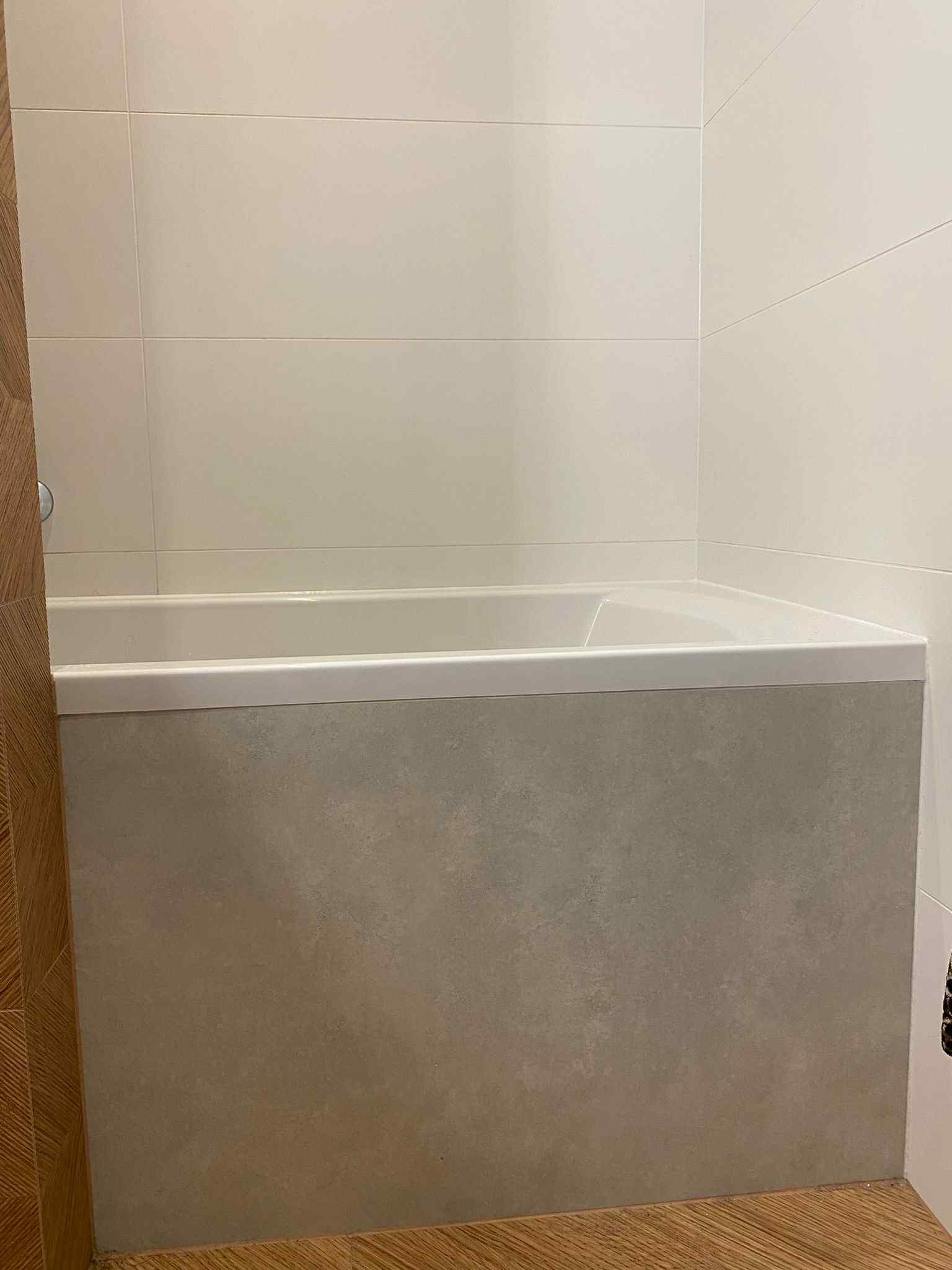 Wykończona wanna z obudową z szarych płytek, białe płytki na ścianie, drewnopodobna podłoga. Minimalistyczny design łazienki po remoncie.