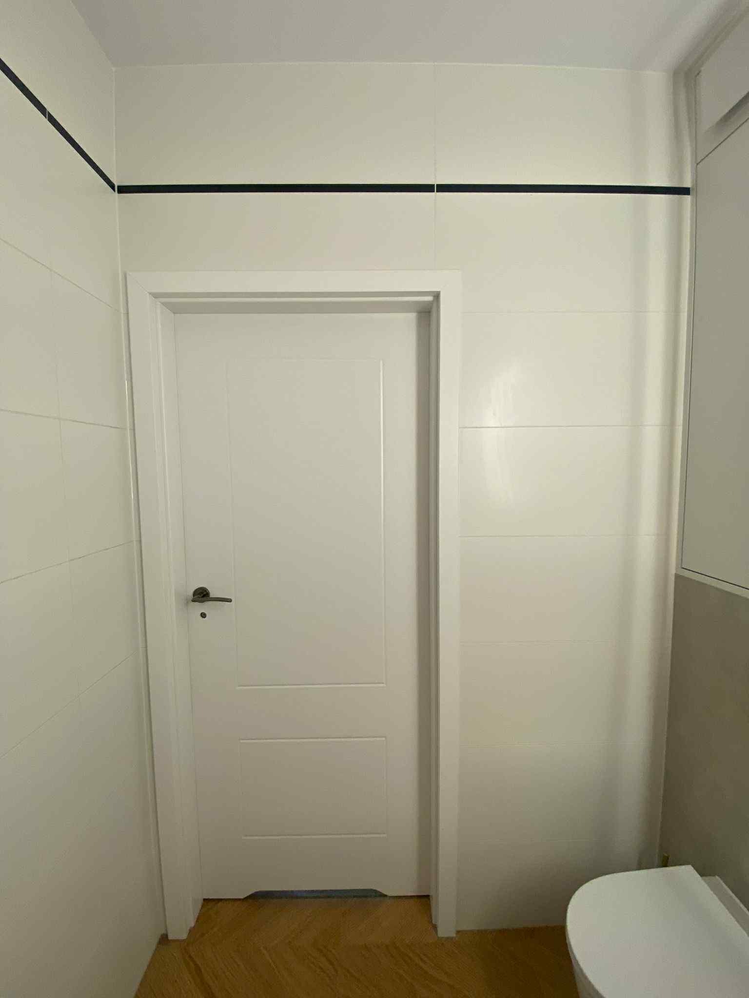 Biała łazienka z jasnymi płytkami i czarnym akcentem w postaci cienkiej linii na górze. Widoczna biała, zamknięta  drzwi i fragment toalety.