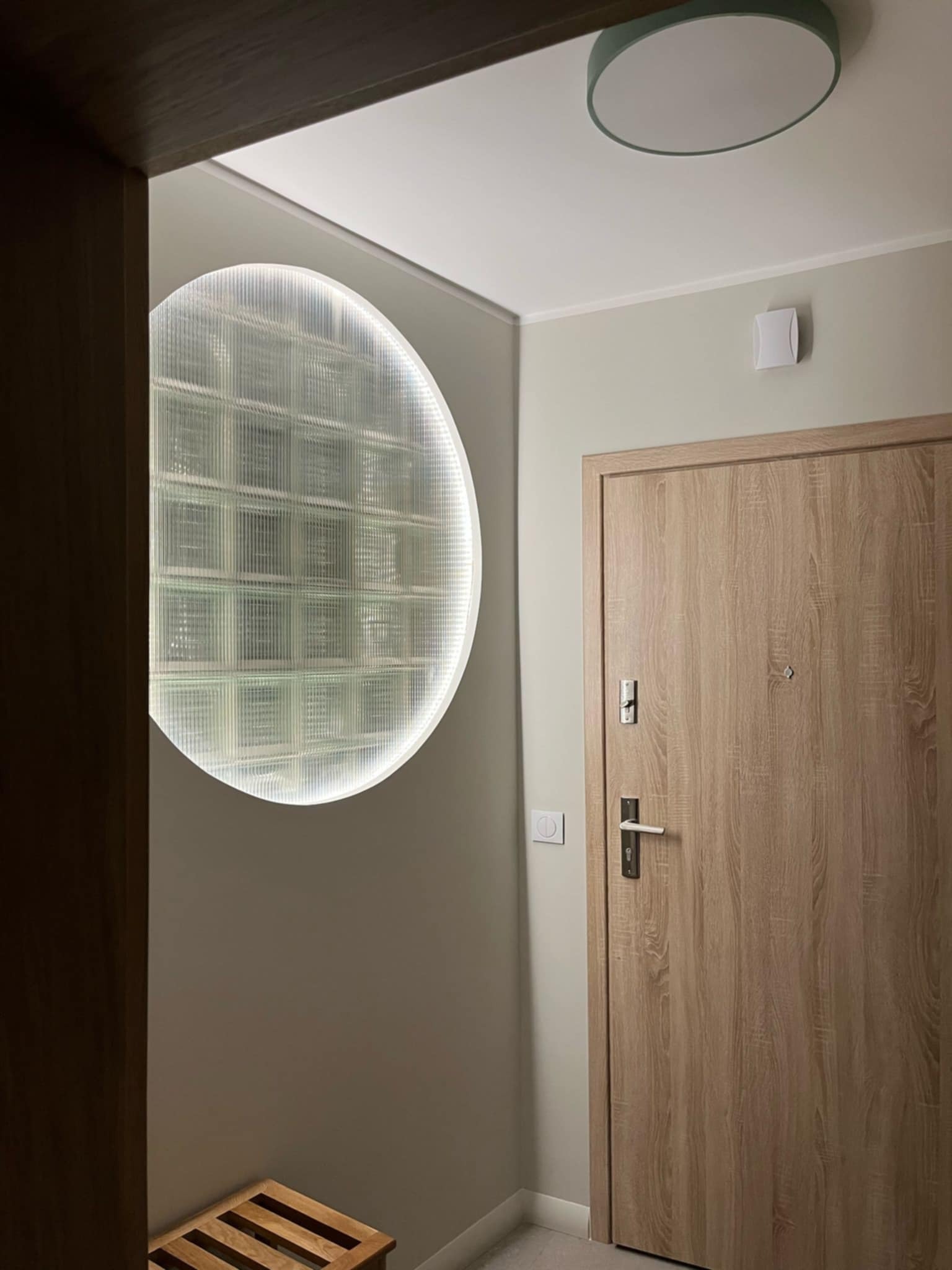 Nowoczesne wnętrze: drewniane drzwi, okrągłe okno z luksferów z podświetleniem LED i minimalistyczna lampa sufitowa. Subtelna kolorystyka ścian.