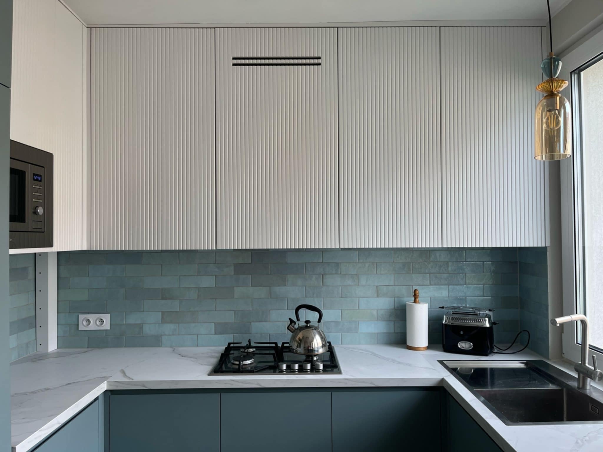 Nowoczesna kuchnia z turkusowymi płytkami na ścianie, białym blatem i szafkami w odcieniach szarości. Widoczny czajnik, toster i designerska lampa.