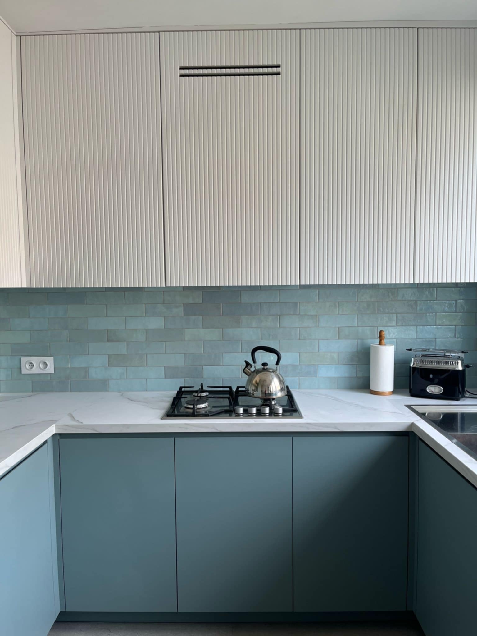 Niebieska zabudowa kuchenna z białym blatem i płytkami w odcieniach błękitu. Na blacie czajnik, toster i akcesoria. Górne szafki białe, ryflowane.