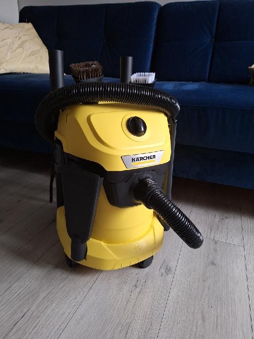 Żółty odkurzacz Karcher z wężem i szczotkami na podłodze, w tle granatowa sofa. Sprzęt do sprzątania po remoncie lub do użytku domowego.