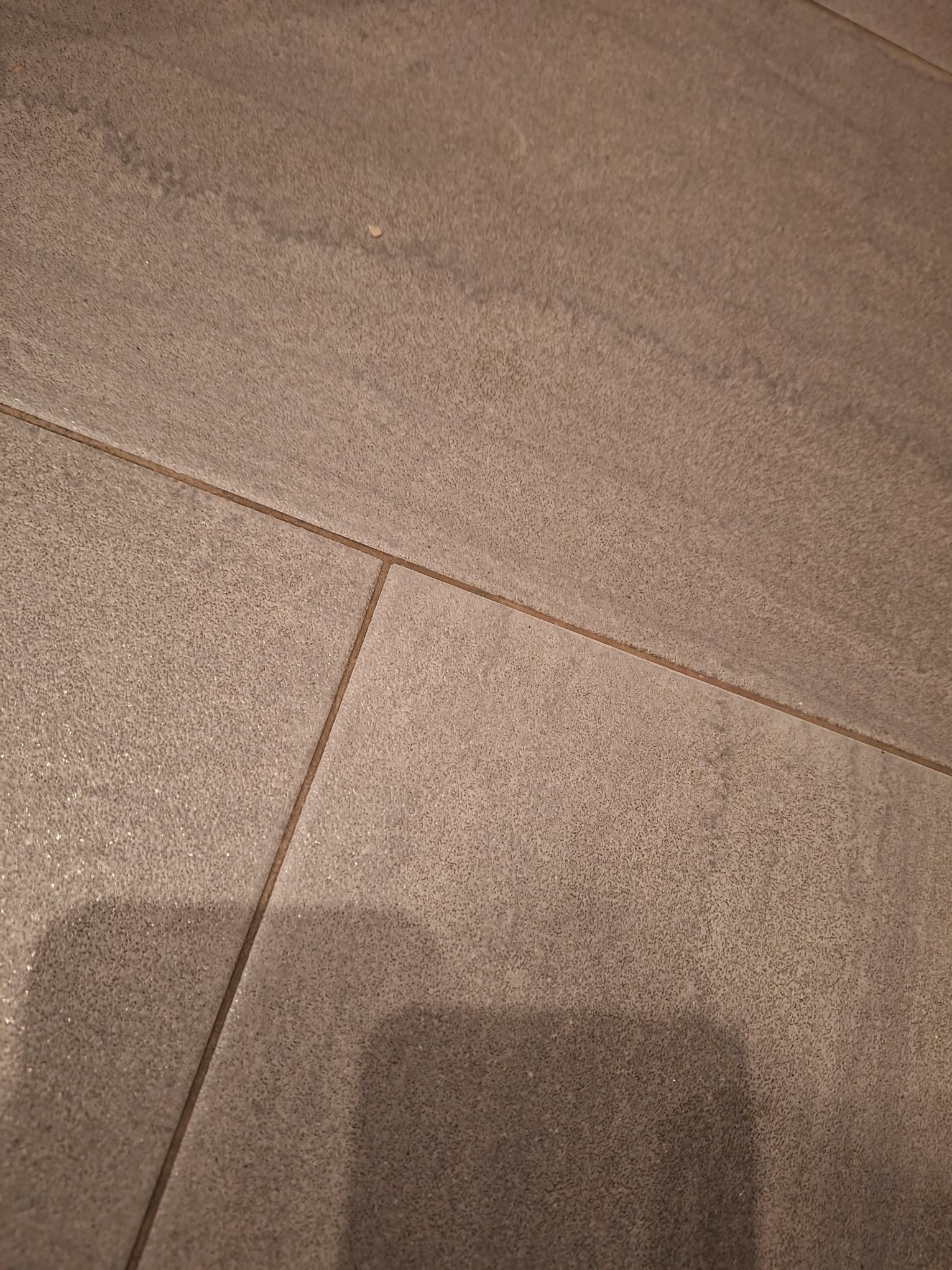 Szare, matowe płytki podłogowe ułożone równo, z widocznymi fugami. Delikatna struktura płytek i ciepłe oświetlenie.