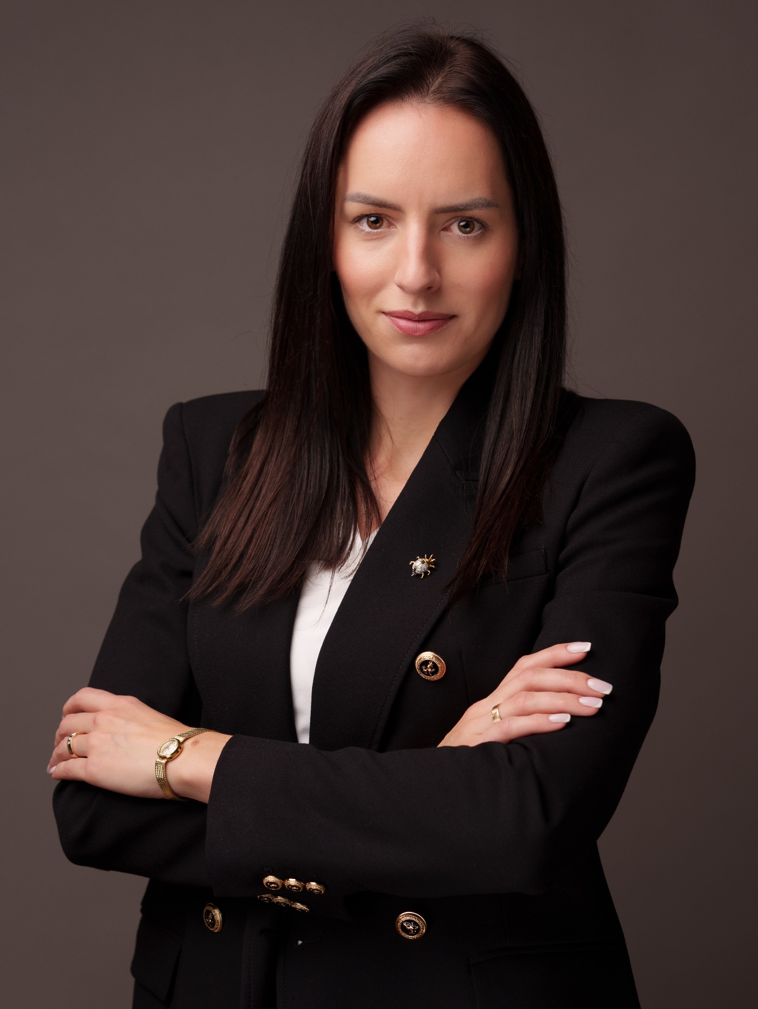 Portret kobiety w czarnej marynarce z broszką i złotym zegarkiem, skrzyżowane ręce, na neutralnym tle. Profesjonalny wizerunek biznesowy.