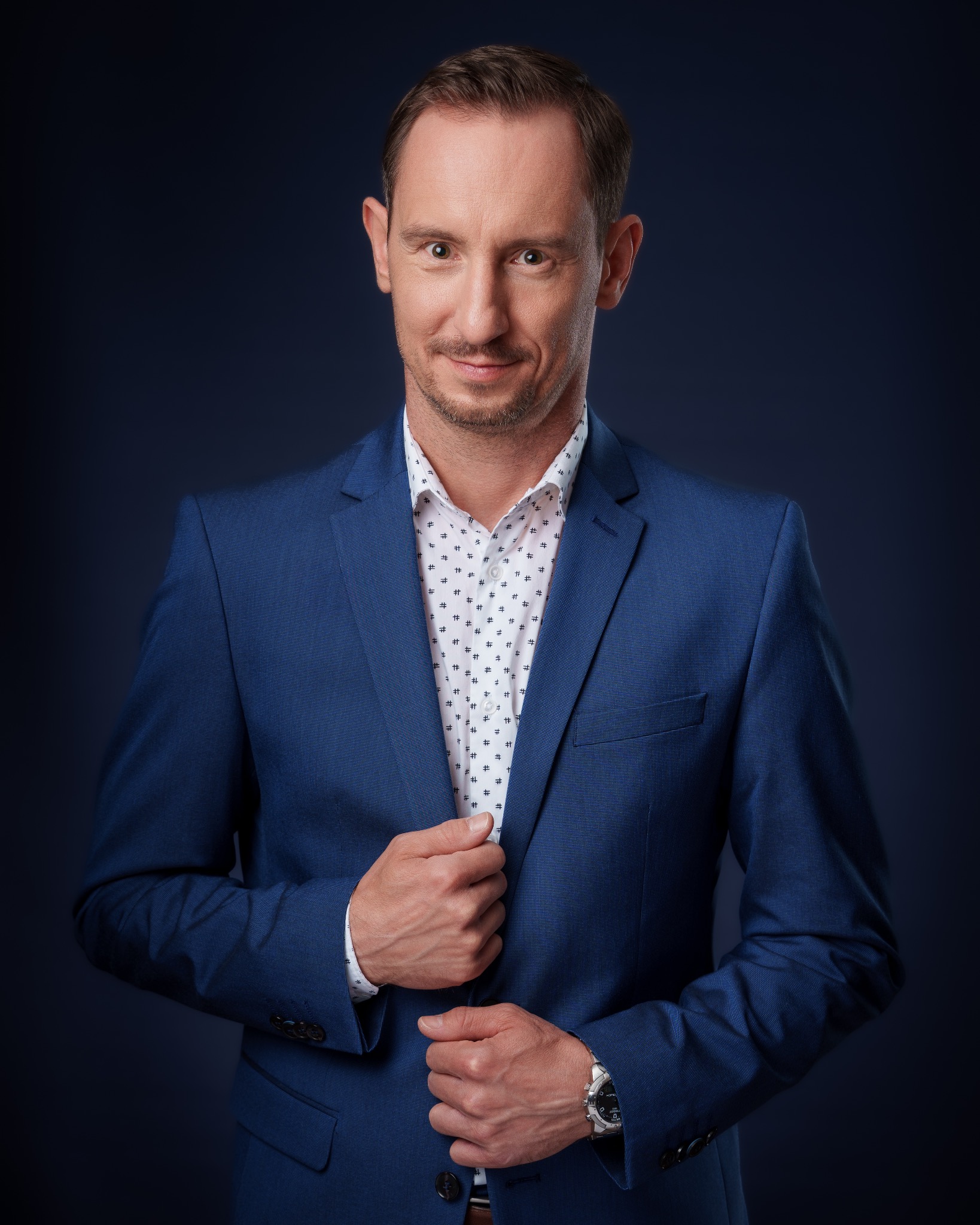 Portret mężczyzny w błękitnym garniturze na ciemnym tle. Uśmiechnięty, z rękami splecionymi na wysokości brzucha. Widoczny zegarek na nadgarstku. Profesjonalny wygląd.