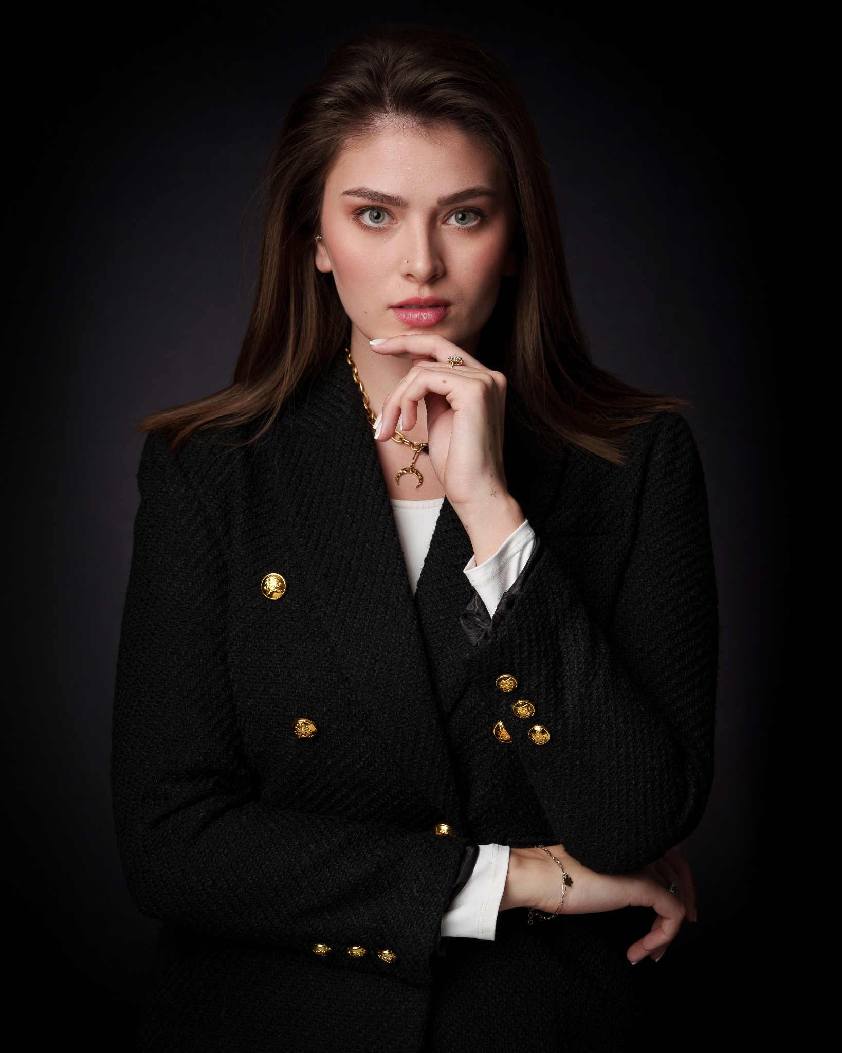 Portret kobiety w czarnym żakiecie z ozdobnymi guzikami, złoty łańcuszek i pierścionek, dłoń podparta pod brodą, ciemne tło, profesjonalna sesja biznesowa.