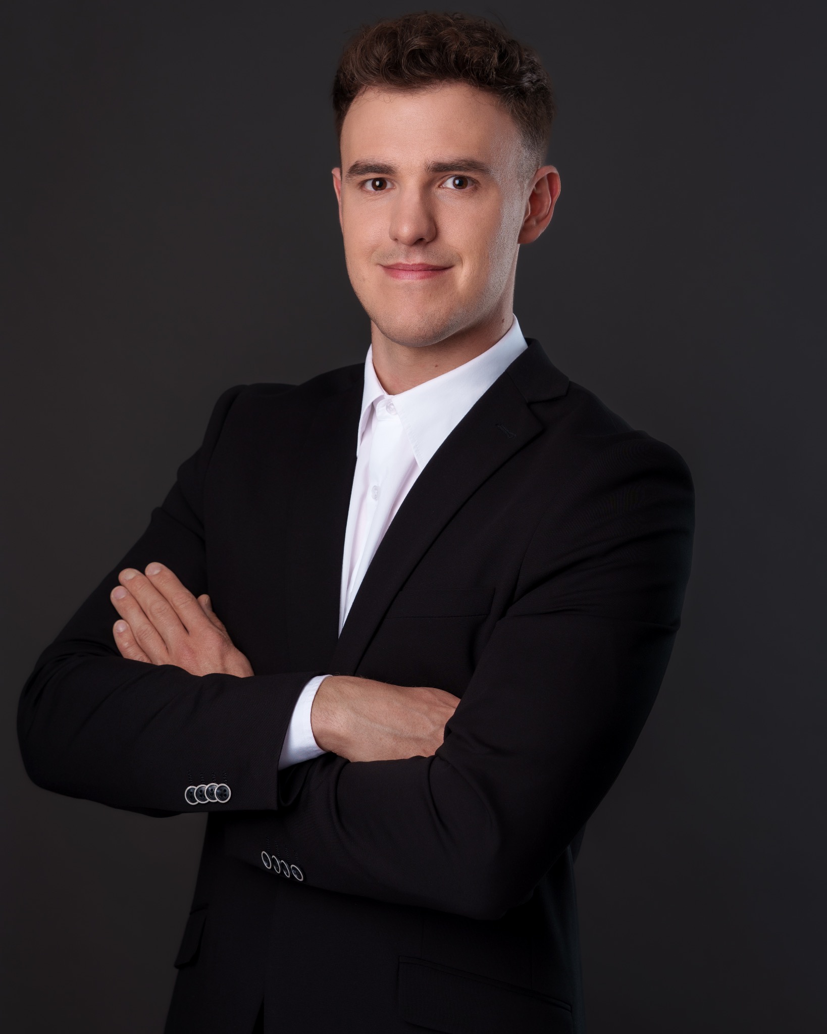 Młody mężczyzna w czarnym garniturze i białej koszuli, z założonymi rękami, uśmiecha się na ciemnoszarym tle. Profesjonalny portret biznesowy.