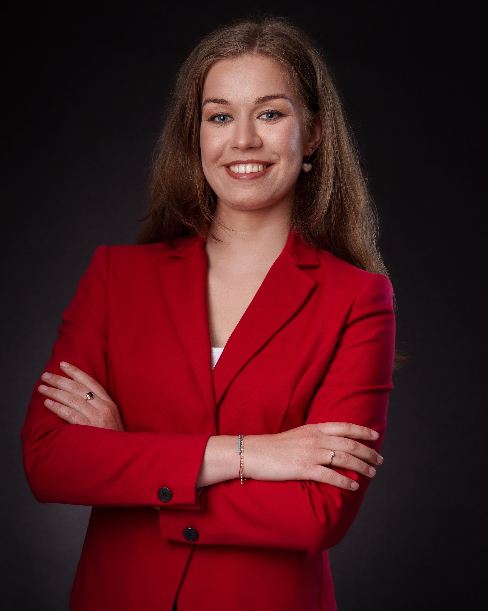 Portret uśmiechniętej kobiety w czerwonej marynarce na ciemnym tle. Skrzyżowane ręce, widoczne pierścionki i bransoletka. Profesjonalny wizerunek biznesowy.