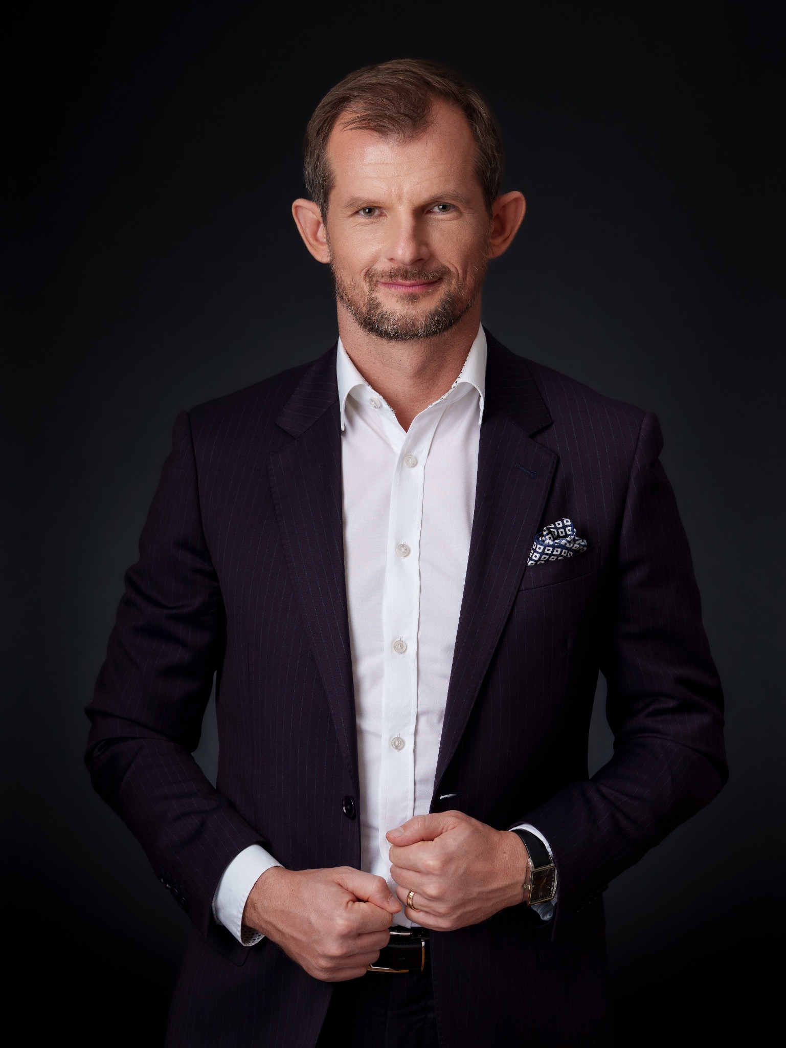 Portret mężczyzny w ciemnym garniturze w prążki i białej koszuli, z poszetką w kieszonce. Uśmiechnięty, z delikatnym zarostem, na ciemnym tle. Widoczny zegarek i obrączka.