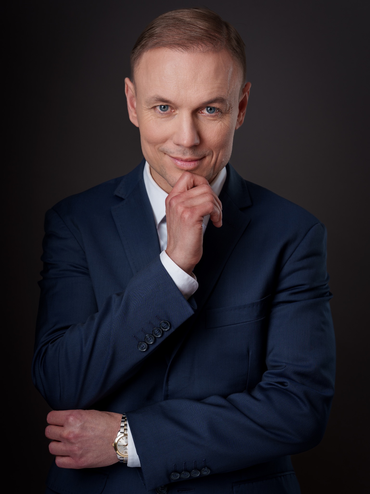 Portret biznesowy mężczyzny w granatowym garniturze na ciemnym tle. Uśmiechnięty, z ręką pod brodą, prezentuje profesjonalny wizerunek.