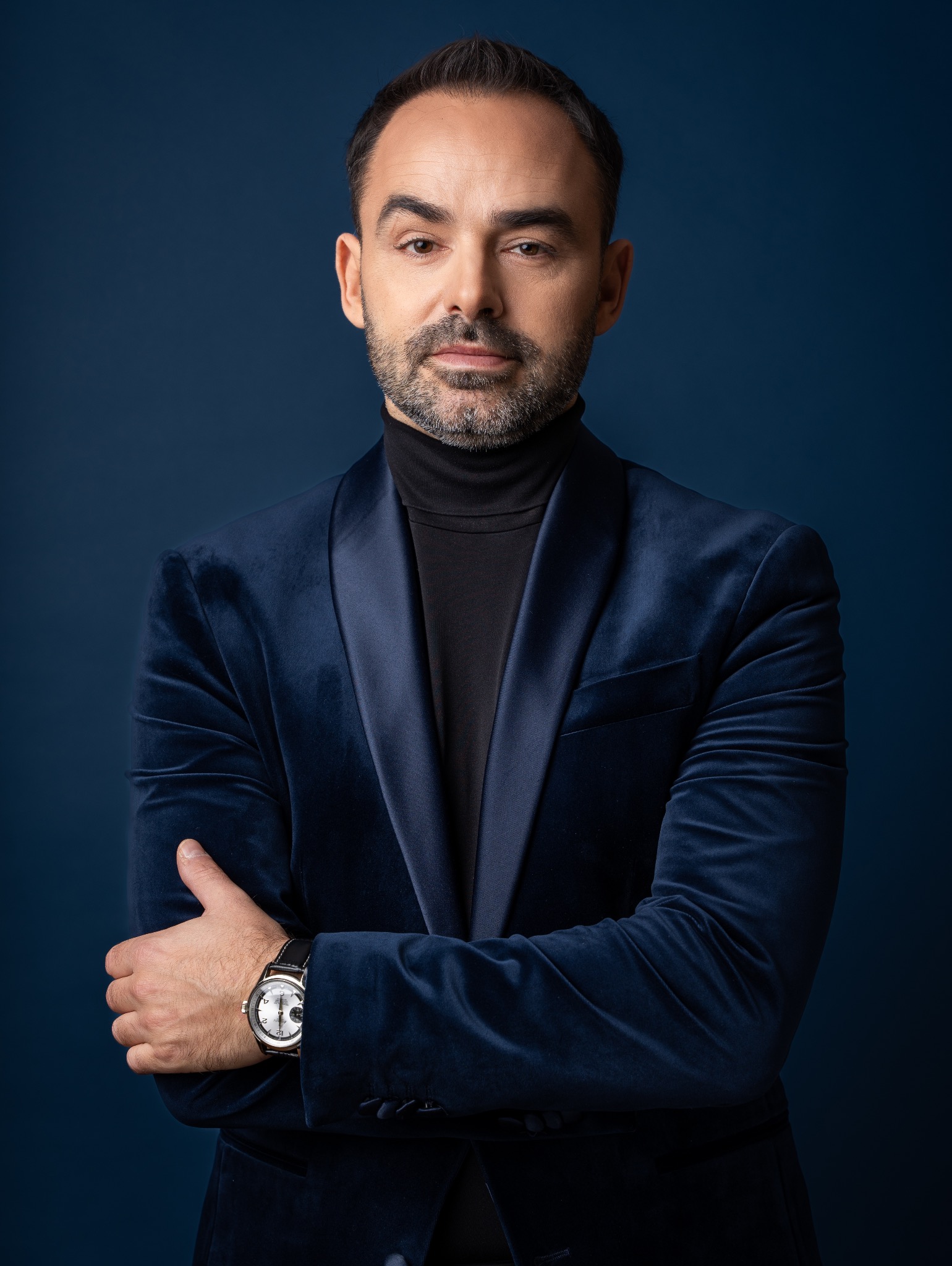 Mężczyzna w aksamitnym, granatowym blezerze i czarnym golfie, z zegarkiem na nadgarstku, pozuje na ciemnoniebieskim tle. Profesjonalny portret biznesowy.