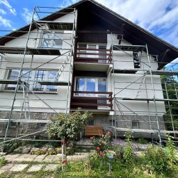 Patryk Kuś PartBud Usługi Budowlane - Dom jednorodzinny w trakcie prac elewacyjnych, otoczony rusztowaniem. Widoczne fragmenty elewacji w jasnym kolorze i kamienne wykończenie. Na pierwszym planie ogród.