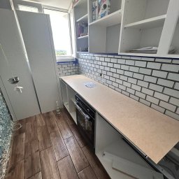 Patryk Kuś PartBud Usługi Budowlane - Montaż kuchni na balkonie: białe szafki wiszące i stojące, brązowe płytki podłogowe imitujące drewno, białe płytki na ścianie. Widoczne elementy instalacji oświetleniowej.