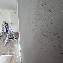 Patryk Kuś PartBud Usługi Budowlane - Ściana po szpachlowaniu z widoczną fakturą, drabina malarska w tle, zabezpieczona podłoga. Prace wykończeniowe w trakcie realizacji.