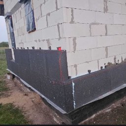 Patryk Kuś PartBud Usługi Budowlane - Ocieplanie fundamentów budynku z bloczków betonowych, widoczne płyty styropianowe mocowane do ściany, teren trawiasty, błękitna beczka w tle.