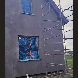 Patryk Kuś PartBud Usługi Budowlane - Szara elewacja domu w trakcie prac wykończeniowych. Okna zabezpieczone folią, obok rusztowanie. Widoczne narzędzia i wiadro. Świeżo nałożony tynk.