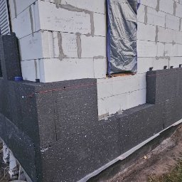 Patryk Kuś PartBud Usługi Budowlane - Ocieplanie ścian zewnętrznych budynku z betonu komórkowego, widoczne płyty styropianowe grafitowe, sznurek traserski i okna zabezpieczone folią.