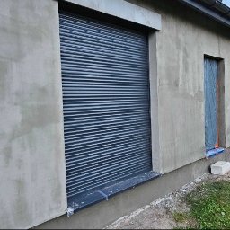 Patryk Kuś PartBud Usługi Budowlane - Budynek po szpachlowaniu z zamontowaną roletą zewnętrzną. Widoczne ślady po pracach wykończeniowych. W tle drewniana altana i zieleń.