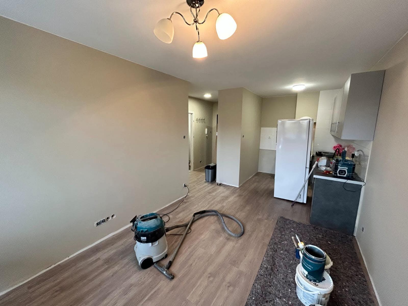 Jasne wnętrze po malowaniu ścian, z widocznym odkurzaczem przemysłowym i farbami. Ściany w odcieniach beżu, podłoga drewnopodobna. Widoczny fragment kuchni.