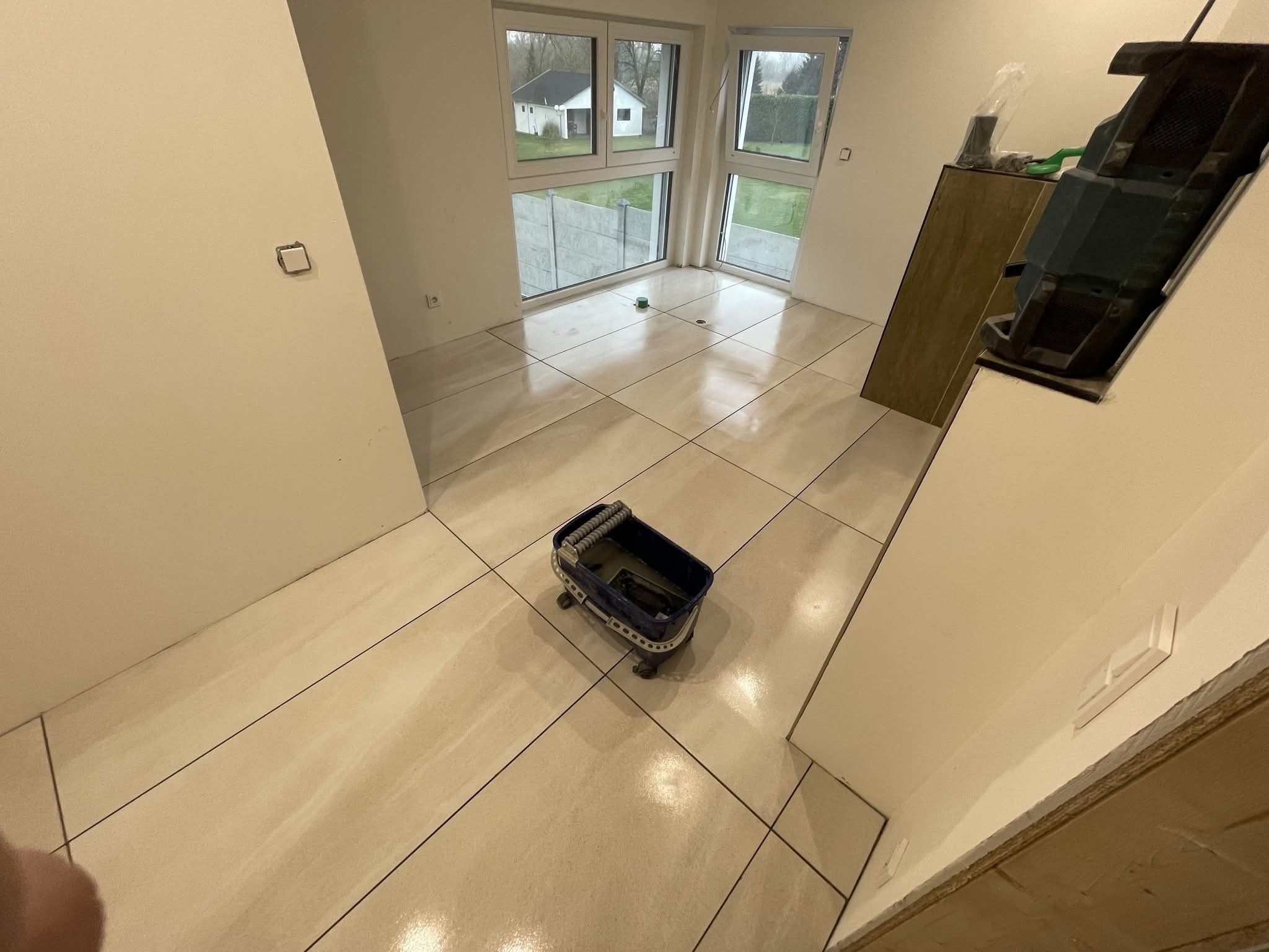 Jasne, kwadratowe płytki podłogowe w świeżo wyremontowanym pokoju z dużymi oknami. Wiadro do mycia płytek na pierwszym planie. Widok na dom z ogrodem za oknem.