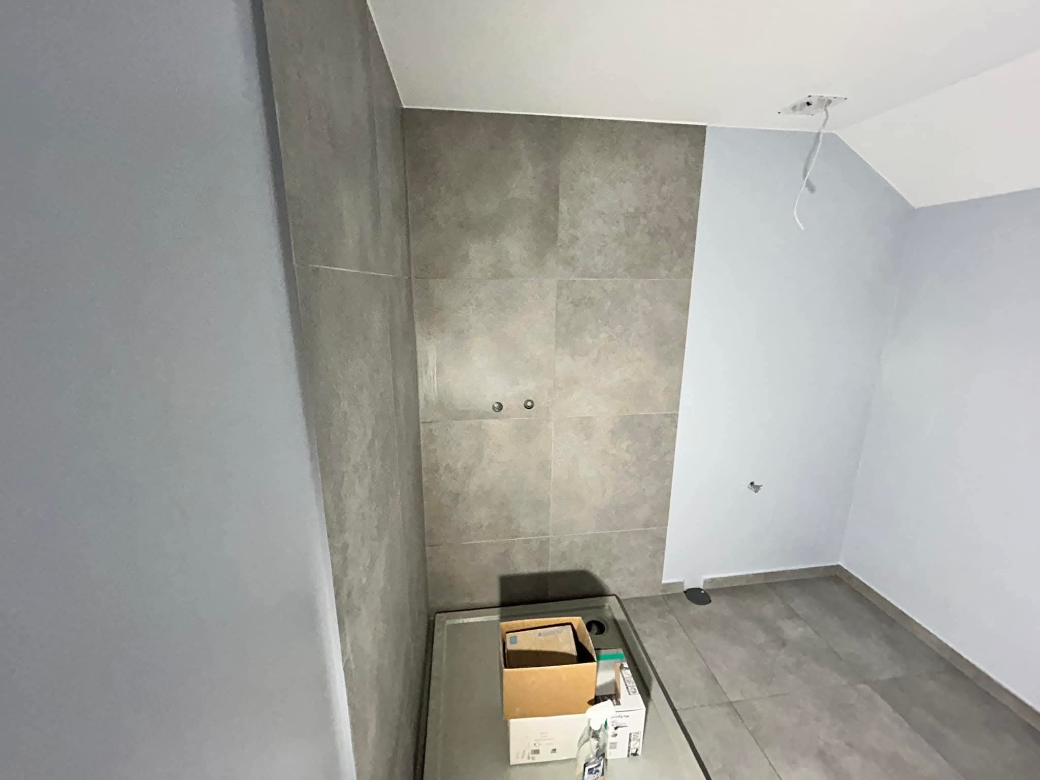 Szara łazienka w trakcie wykańczania: płytki na ścianach, brodzik, kartony z materiałami. Widoczne przygotowania do montażu armatury i oświetlenia.