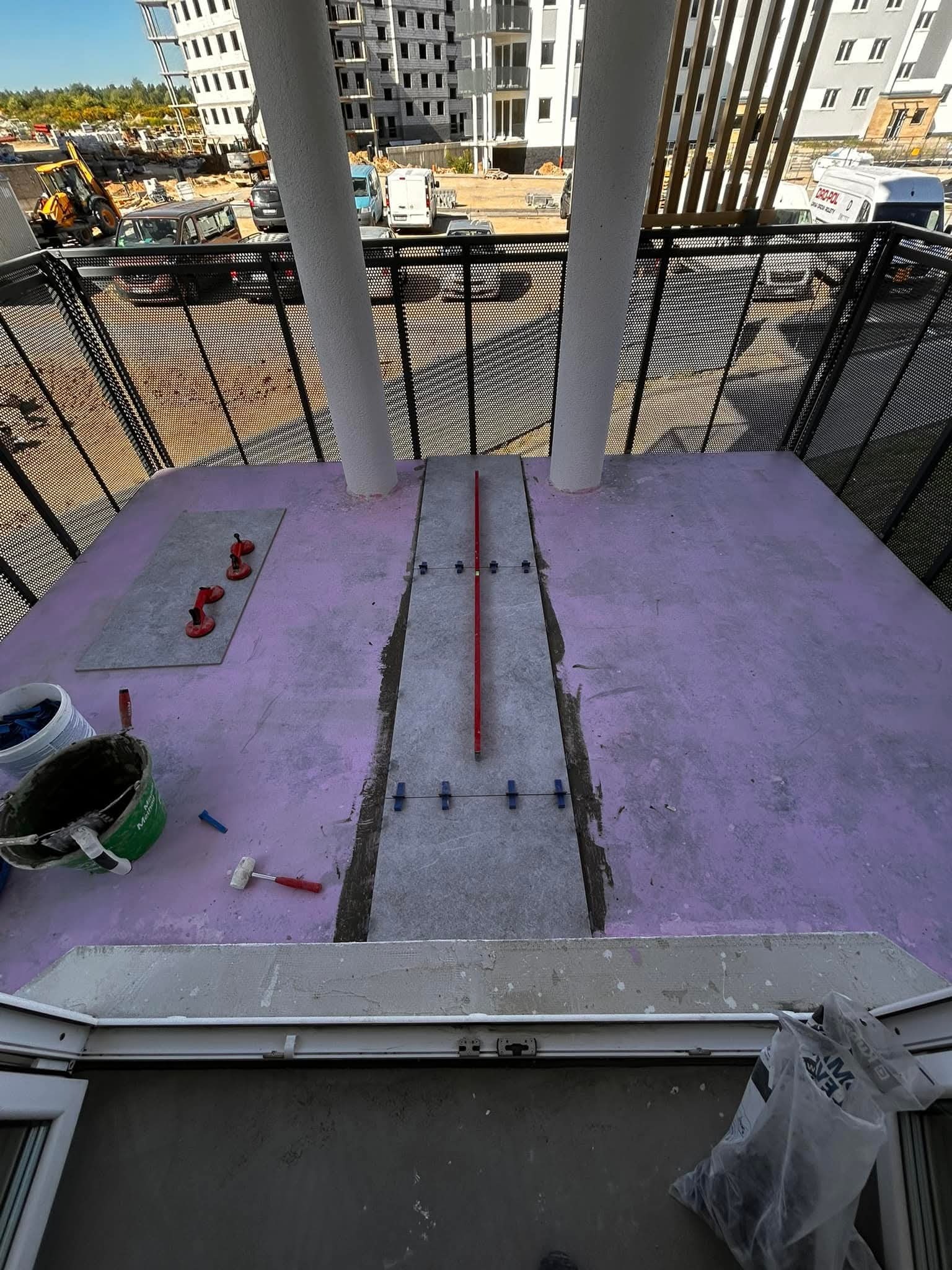 Balkon w trakcie układania płytek podłogowych. Widoczne narzędzia glazurnicze, poziomica i krzyżyki dystansowe. W tle budynek w budowie i metalowa balustrada.