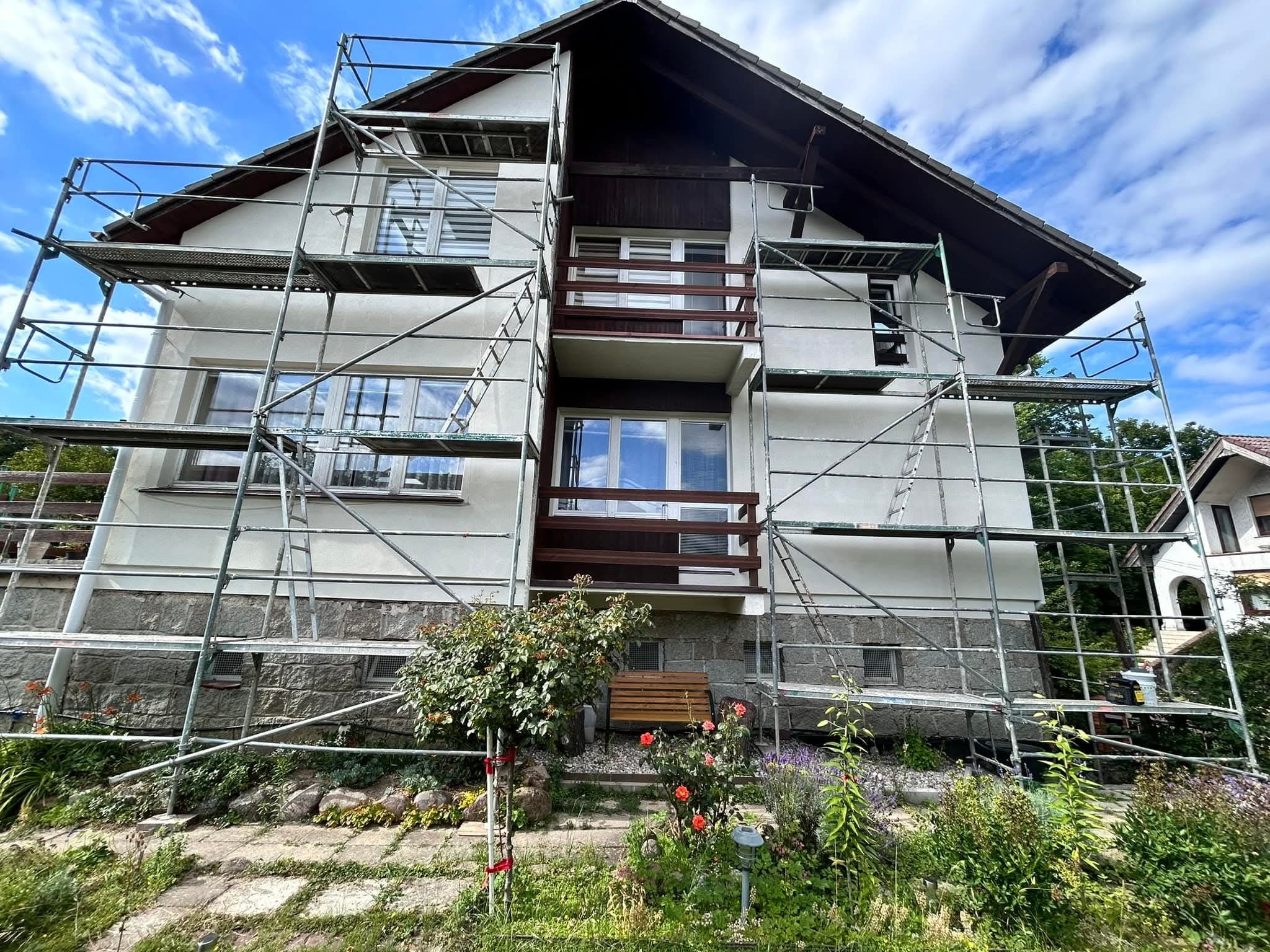 Dom jednorodzinny w trakcie prac elewacyjnych, otoczony rusztowaniem. Widoczne fragmenty elewacji w jasnym kolorze i kamienne wykończenie. Na pierwszym planie ogród.