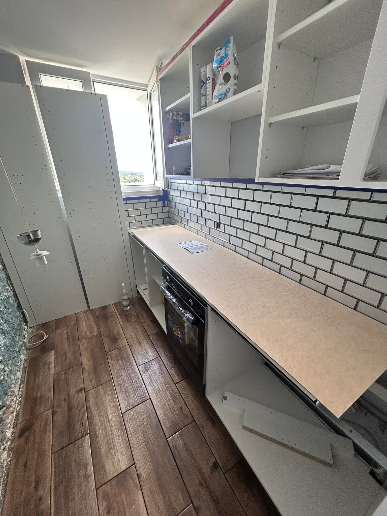 Montaż kuchni na balkonie: białe szafki wiszące i stojące, brązowe płytki podłogowe imitujące drewno, białe płytki na ścianie. Widoczne elementy instalacji oświetleniowej.