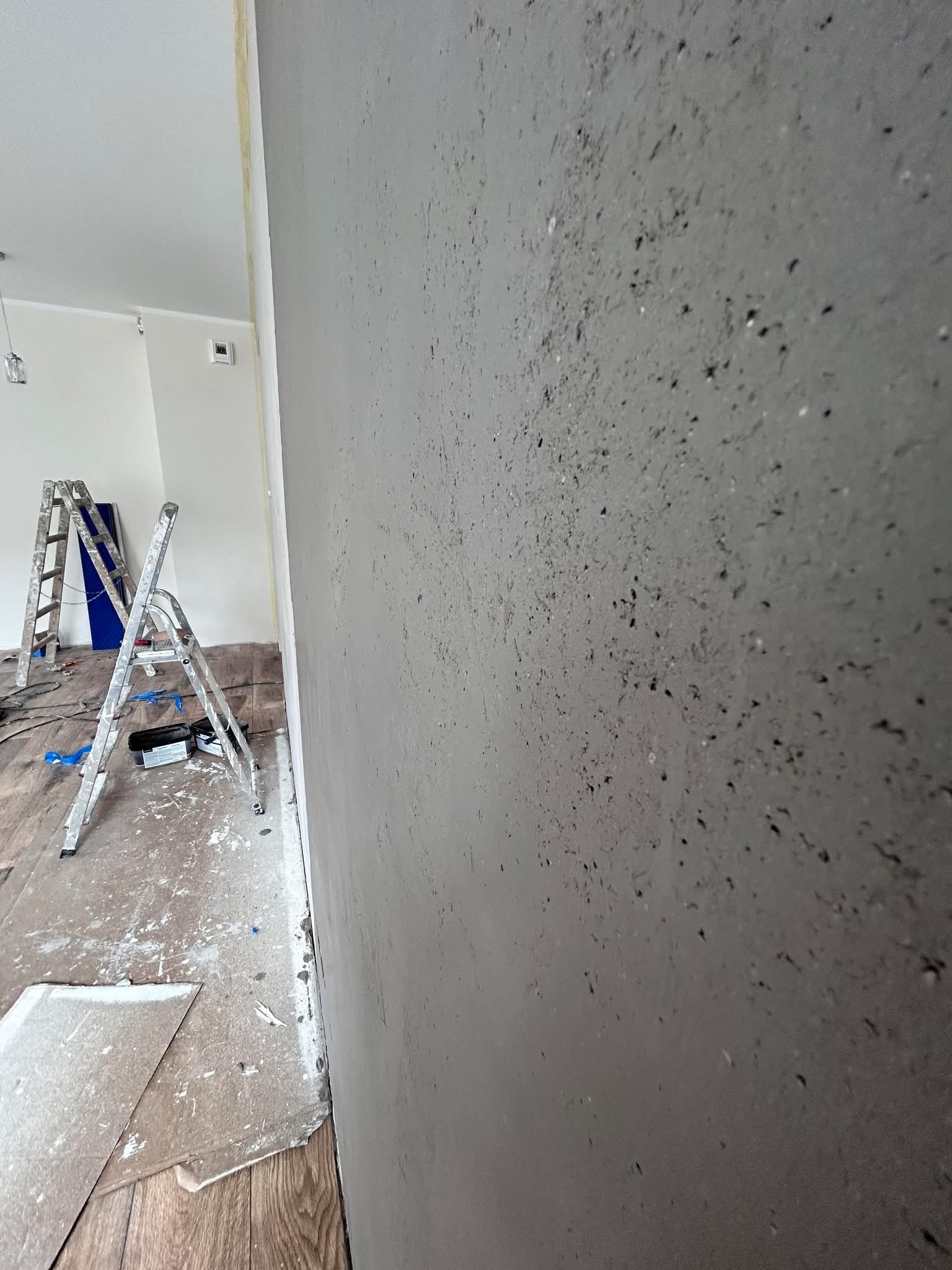 Ściana po szpachlowaniu z widoczną fakturą, drabina malarska w tle, zabezpieczona podłoga. Prace wykończeniowe w trakcie realizacji.