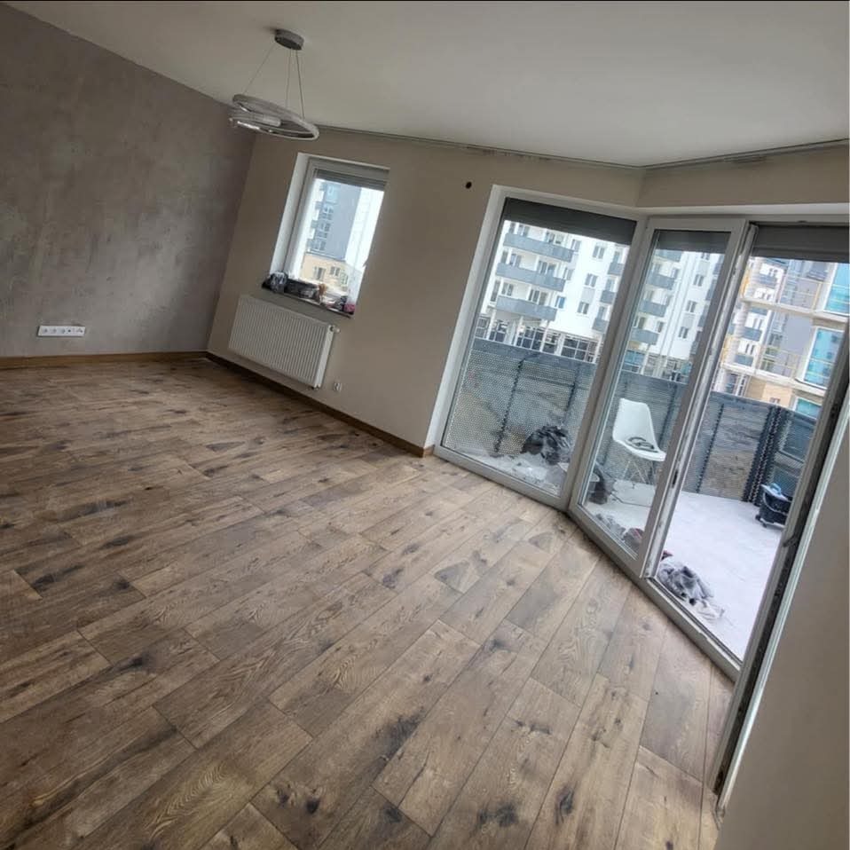 Ukończona podłoga z paneli drewnianych w jasnym pokoju z oknem i wyjściem na balkon. Widoczny grzejnik i nowoczesna lampa sufitowa.