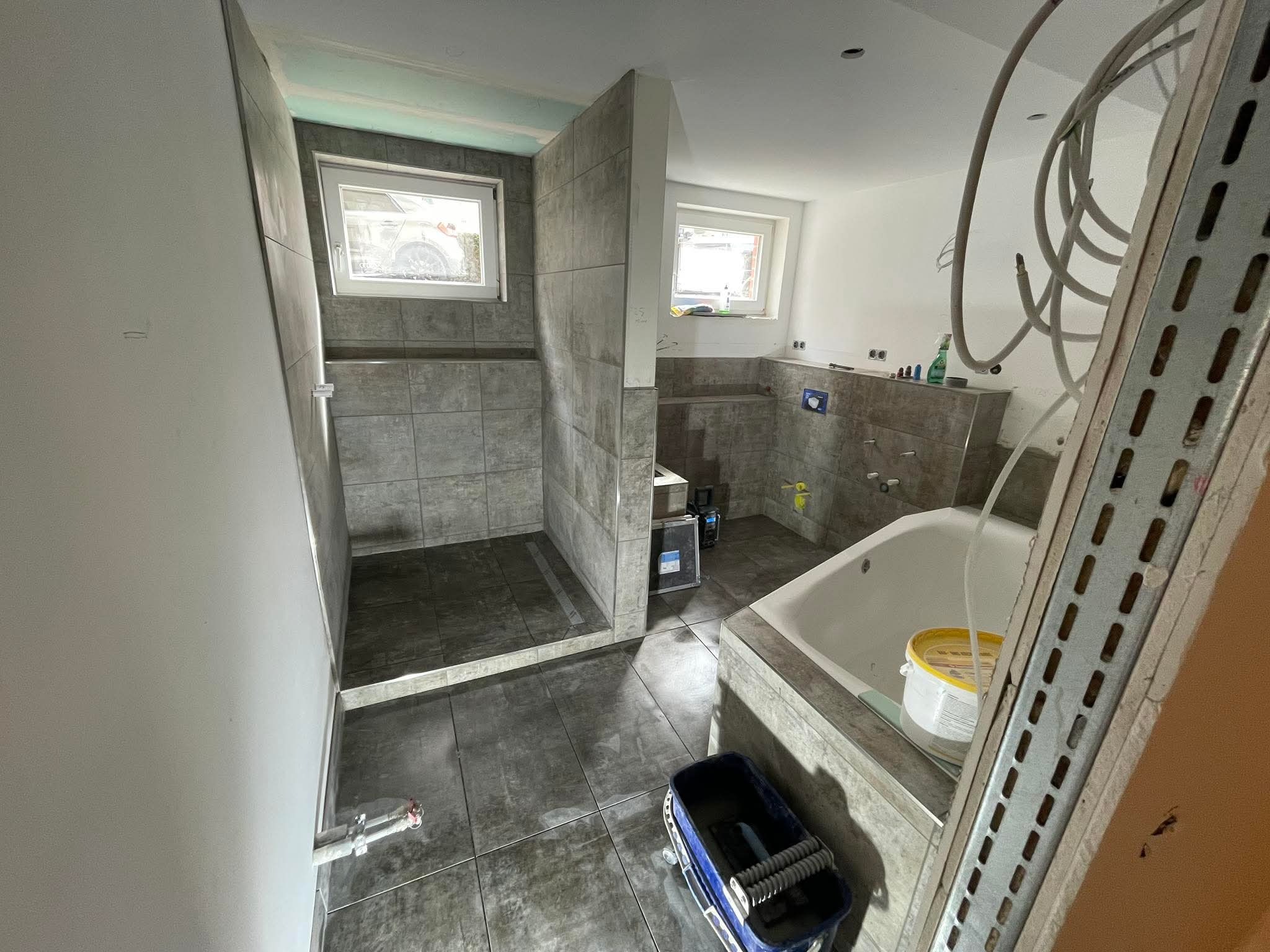 Wnętrze łazienki w trakcie prac wykończeniowych: ułożone płytki podłogowe i ścienne, wanna, widoczne narzędzia glazurnicze i materiały budowlane. Surowy stan, brak armatury.