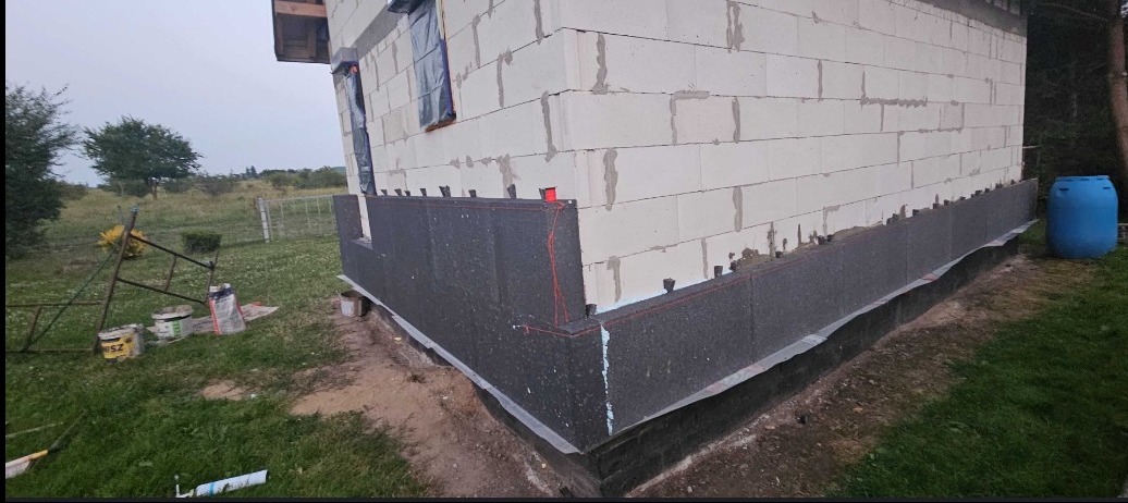 Ocieplanie fundamentów budynku z bloczków betonowych, widoczne płyty styropianowe mocowane do ściany, teren trawiasty, błękitna beczka w tle.